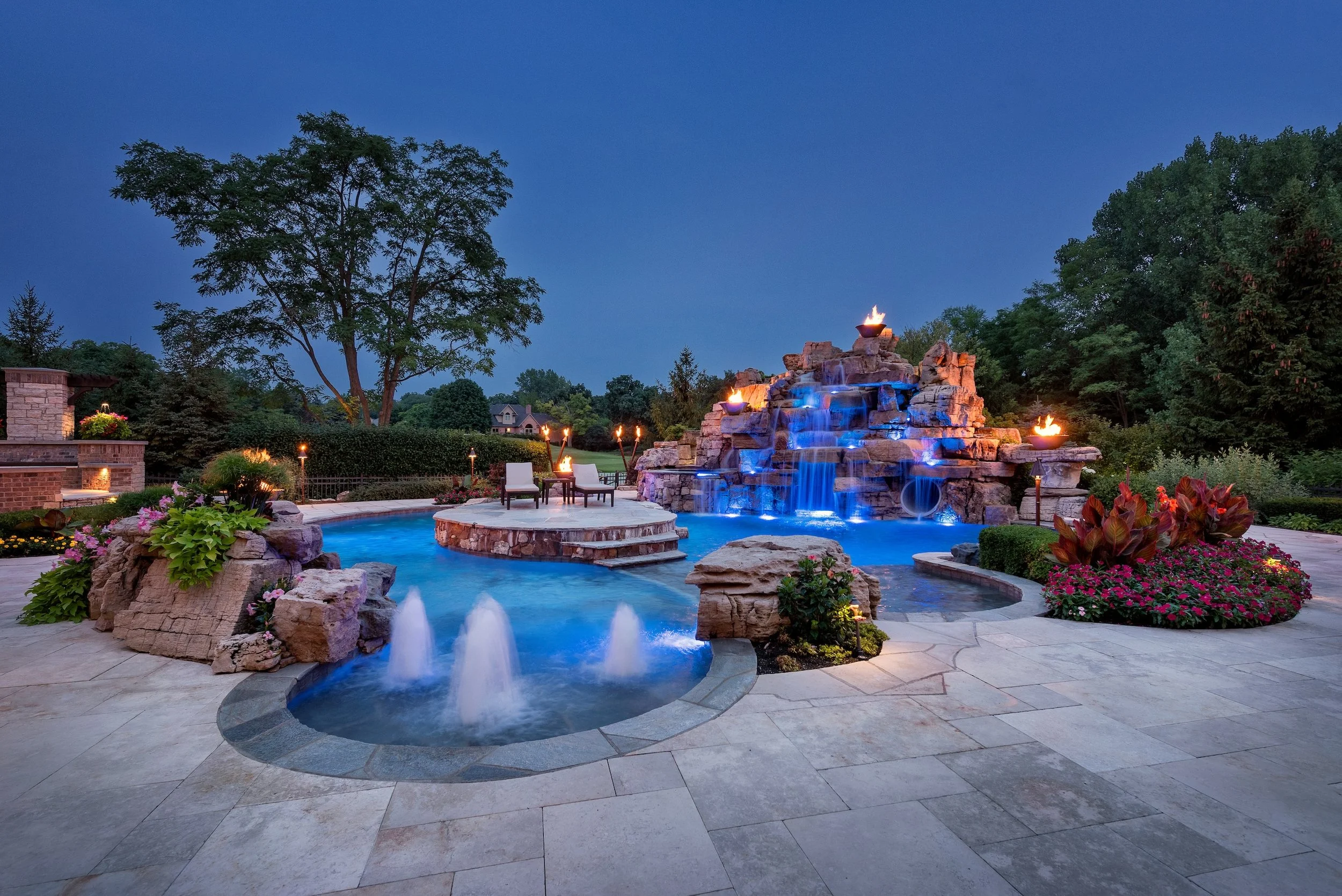 Luxurious Chicago backyard with a swimming pool featuring blue lighting, a waterfall, fire features, a small attached hot tub, and landscaped surroundings with plants and flowers at dusk.