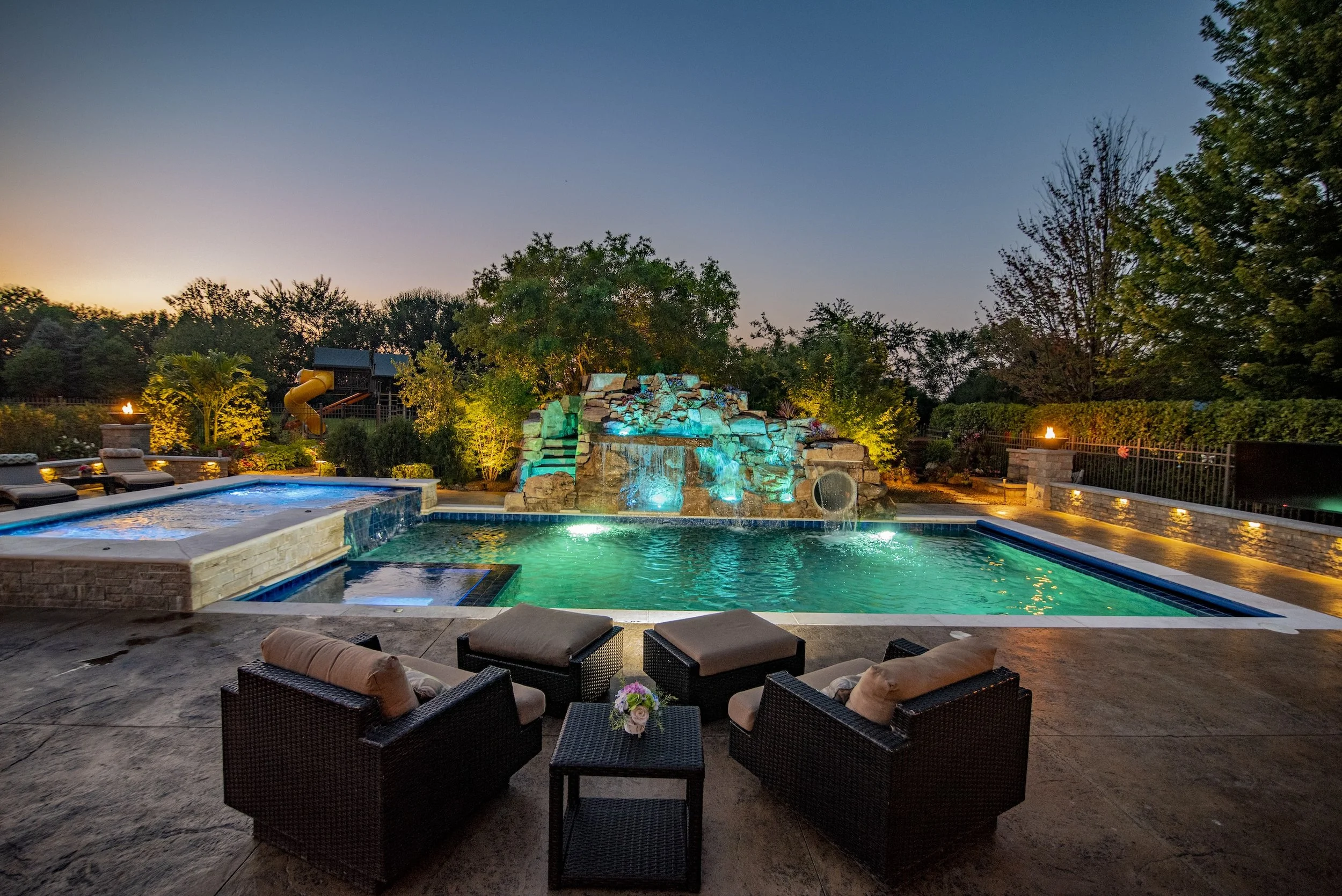 Luxury backyard pool with rock waterfall, illuminated at dusk, outdoor seating, and a background of trees and a slide.