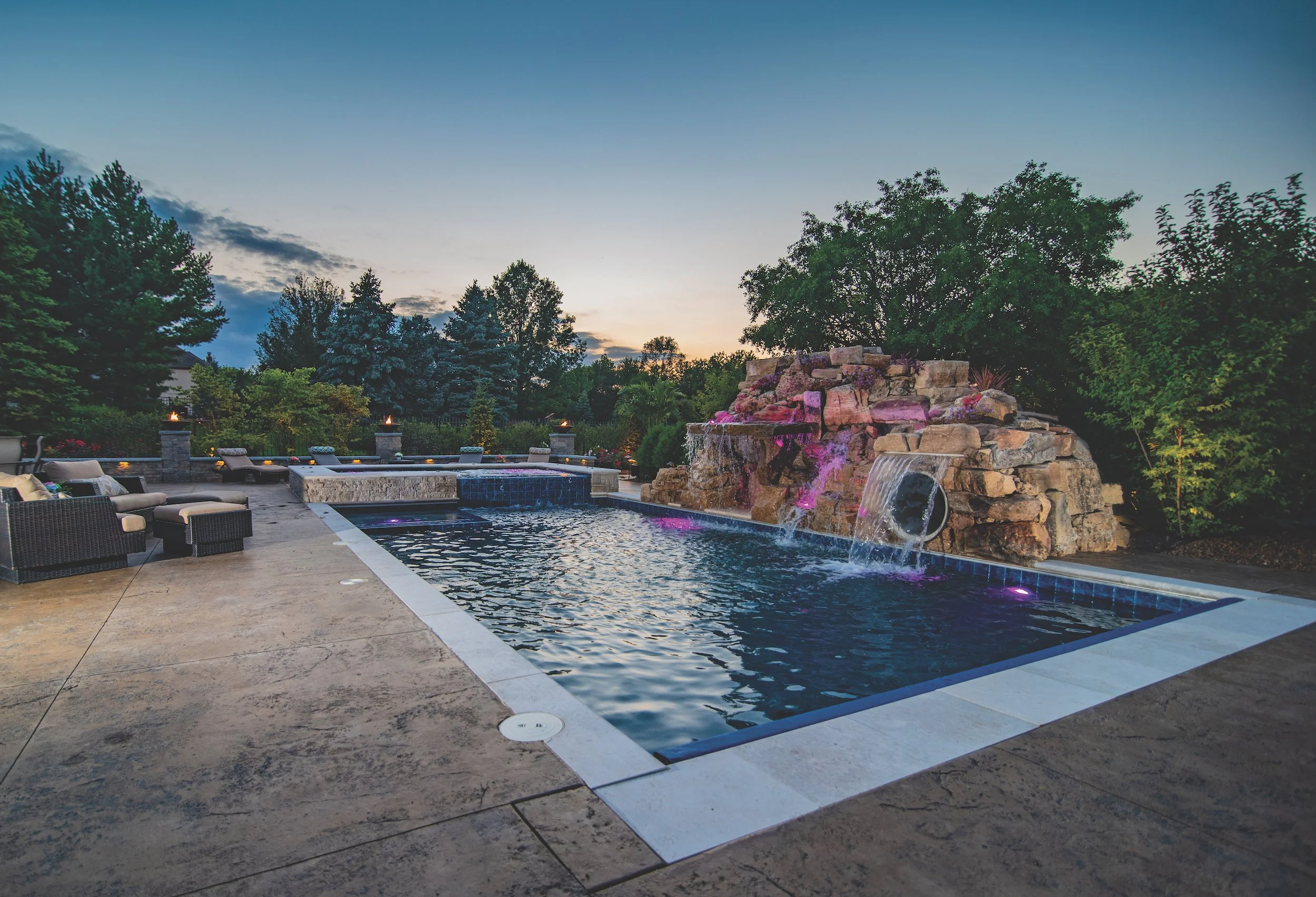 Outdoor swimming pool with waterfall feature surrounded by patio furniture and landscaping at sunset.