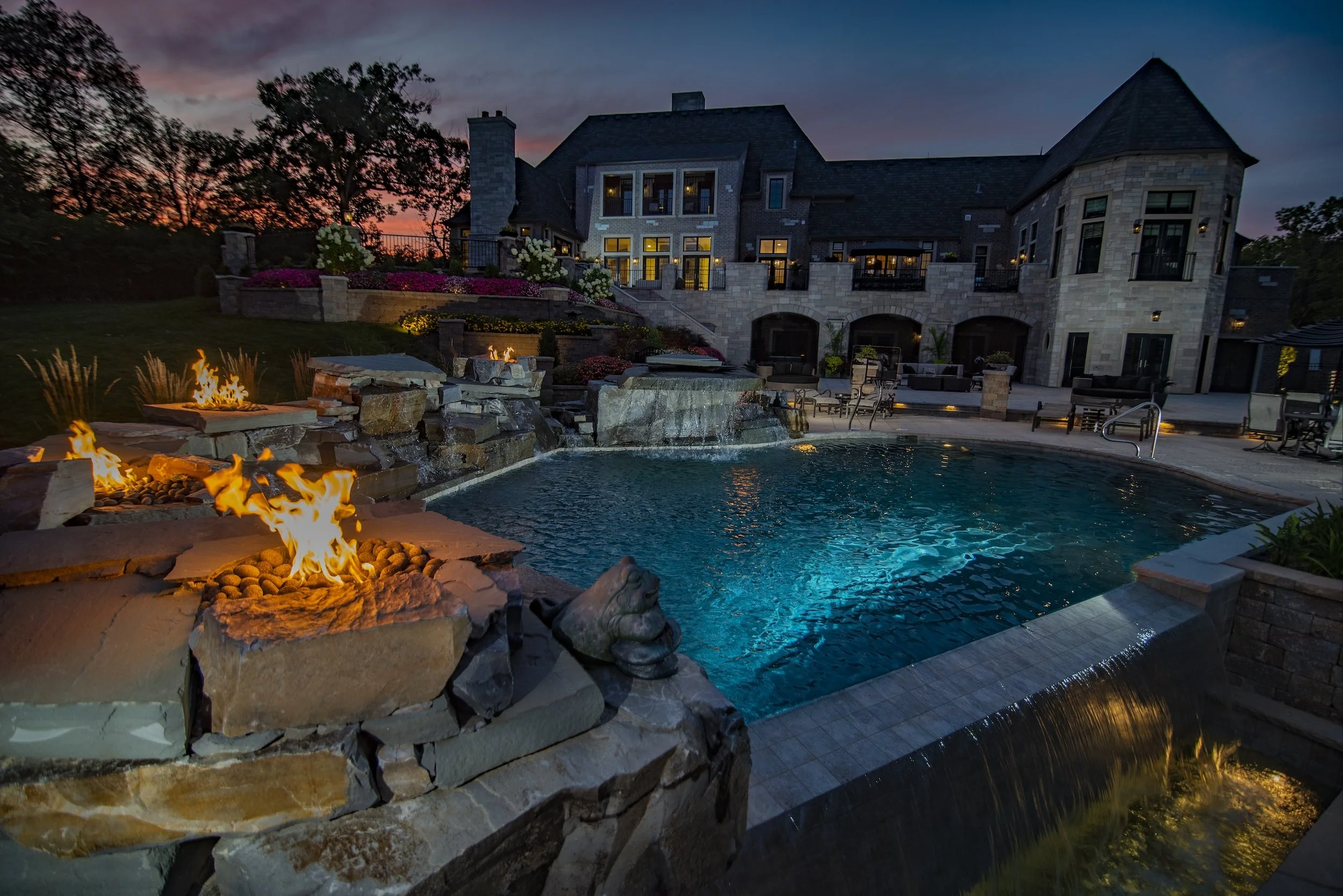 Luxurious backyard with a large pool and fire pits, adjacent to a grand modern mansion at dusk.