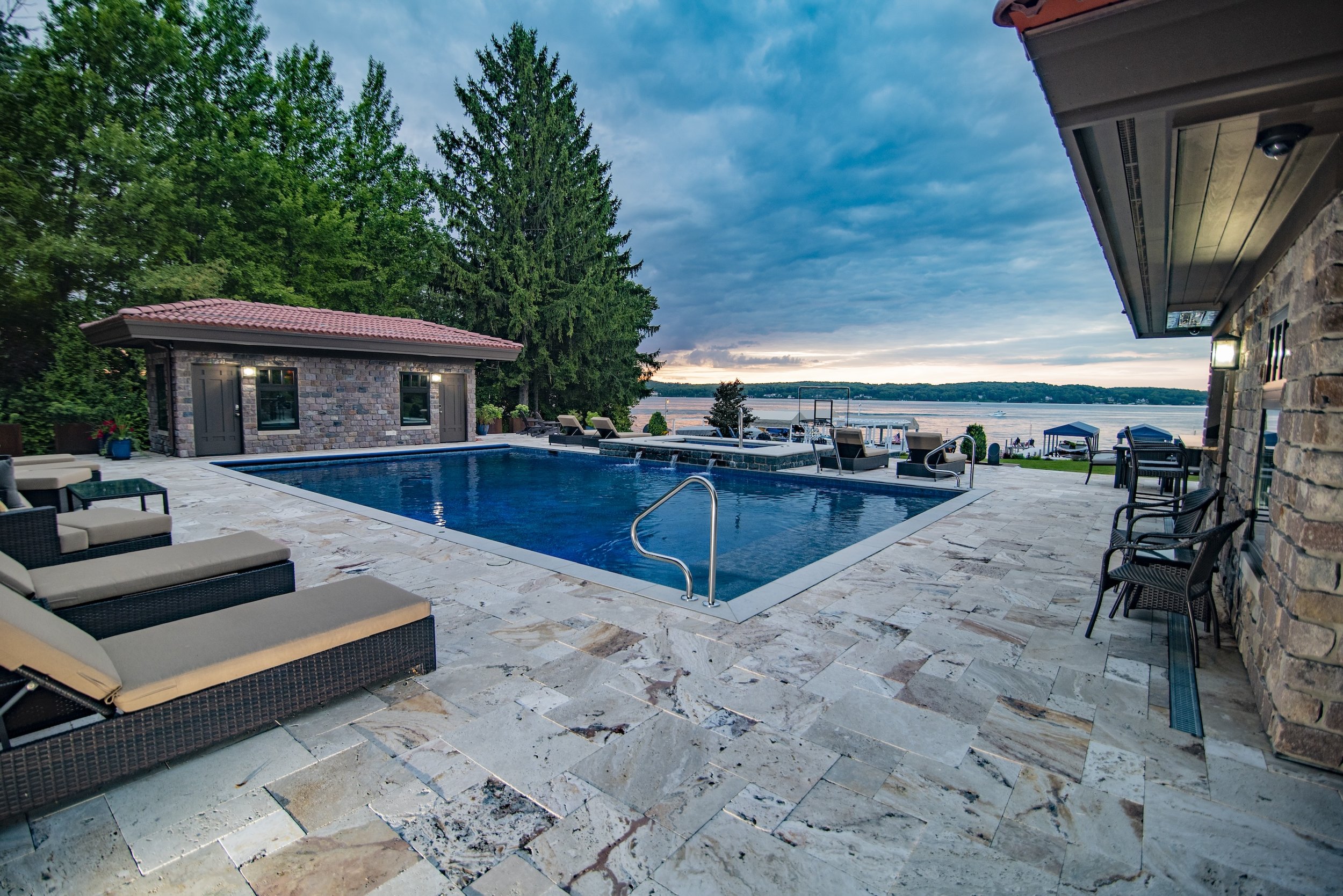 Luxury poolside with lounge chairs, stone patio, small pool house, overlooking a lake with boats, surrounded by trees.