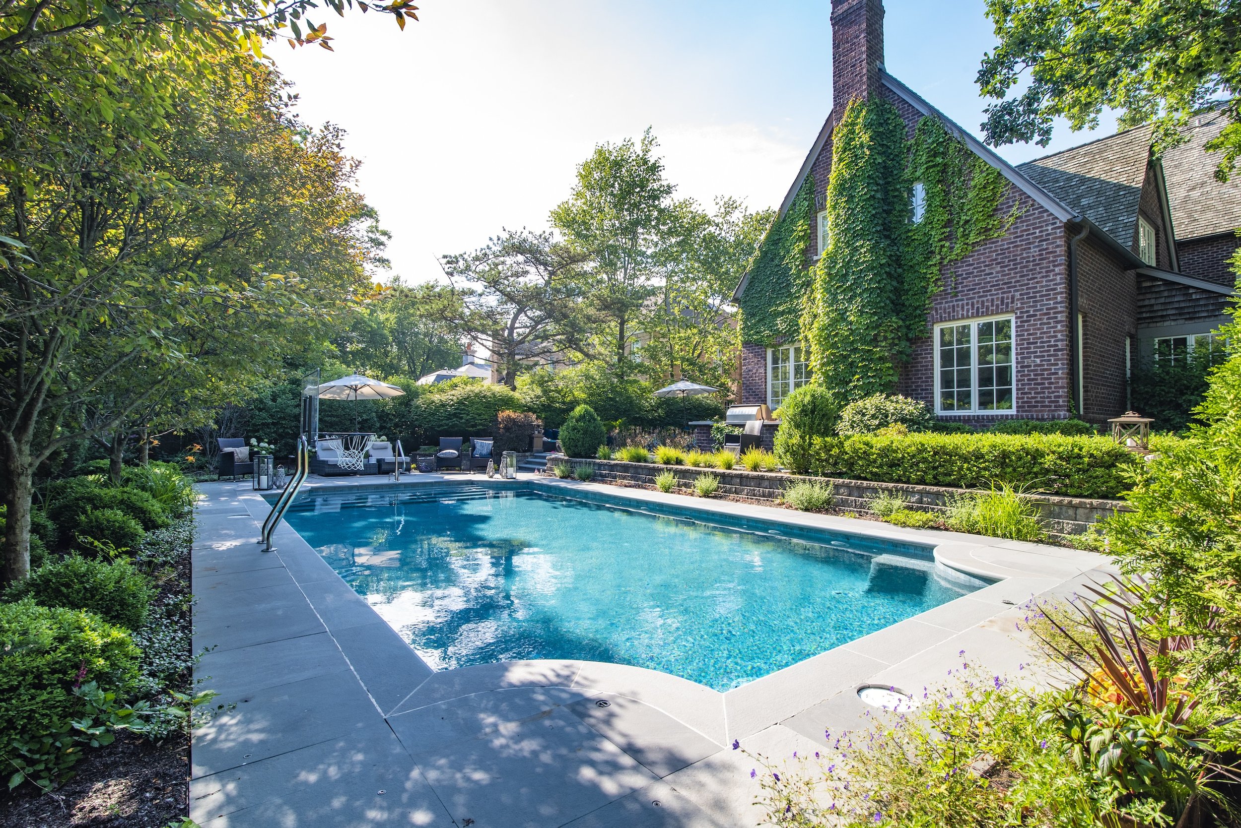 Backyard with rectangular swimming pool, surrounded by trees and shrubs, next to a brick house with ivy on the walls.