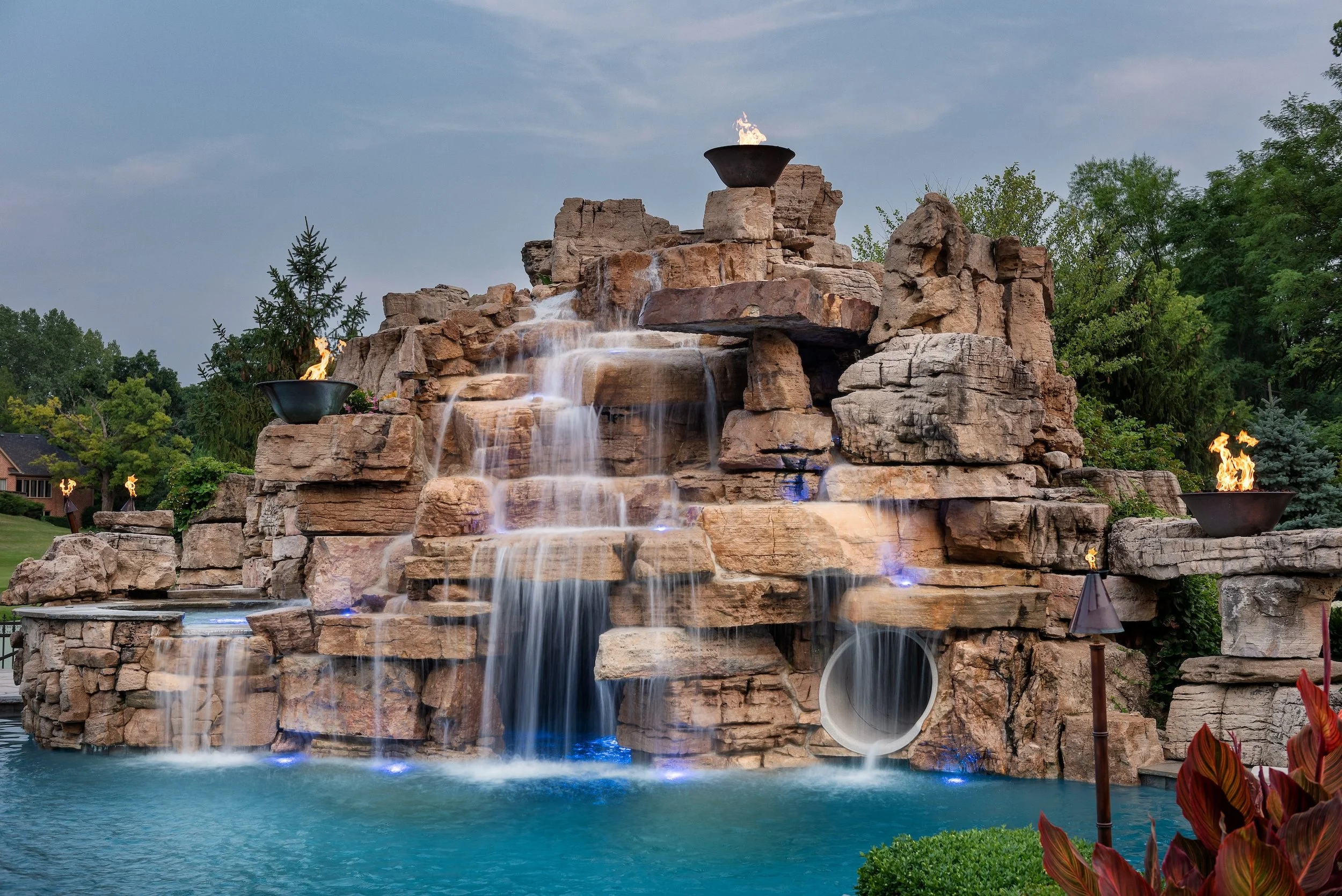 Large rock waterfall structure with flowing water and fire pits on top, surrounded by greenery.