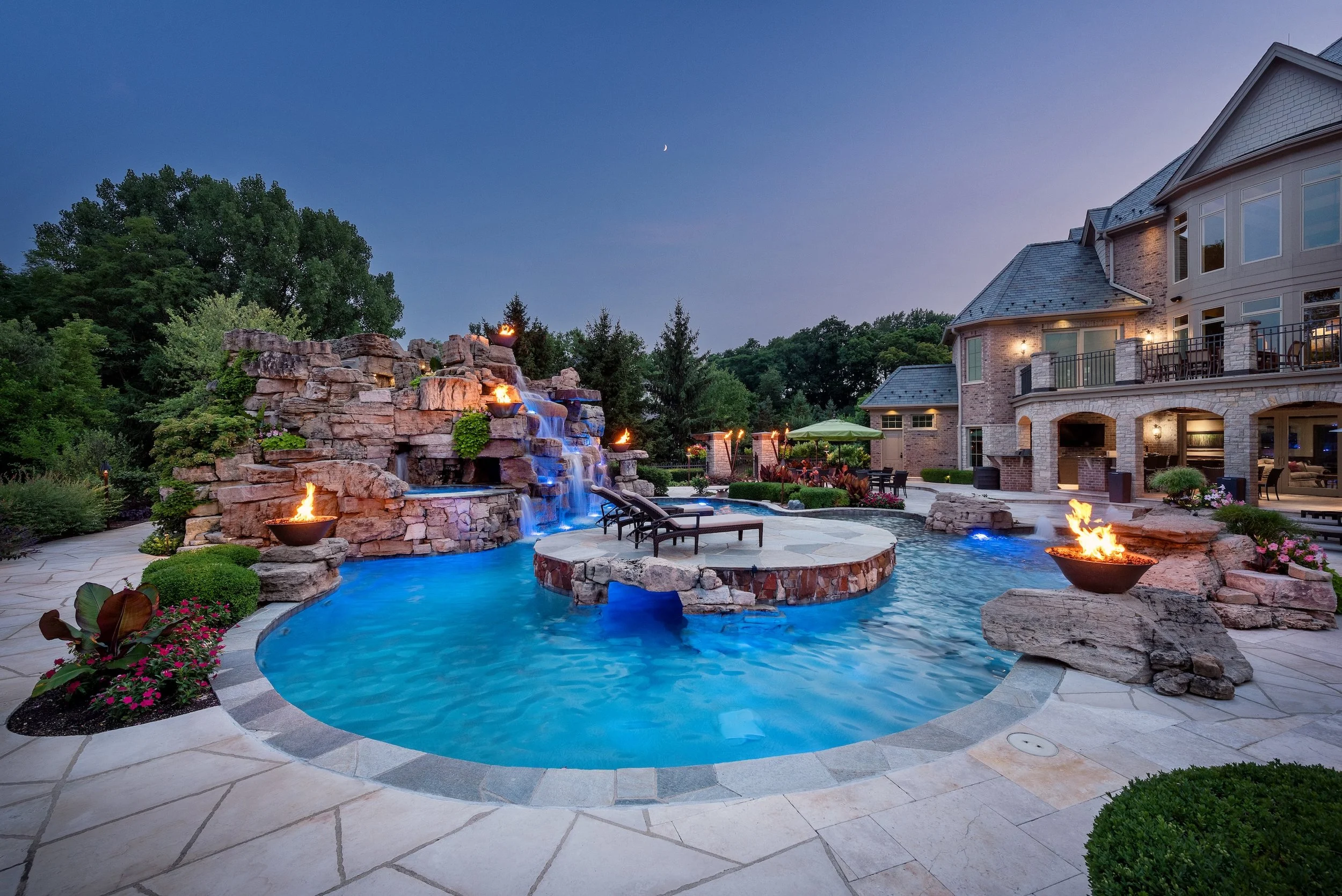 Luxurious outdoor pool area with a large waterfall feature and fire bowls, surrounded by lush landscaping and a stone patio, adjacent to an upscale house during dusk.