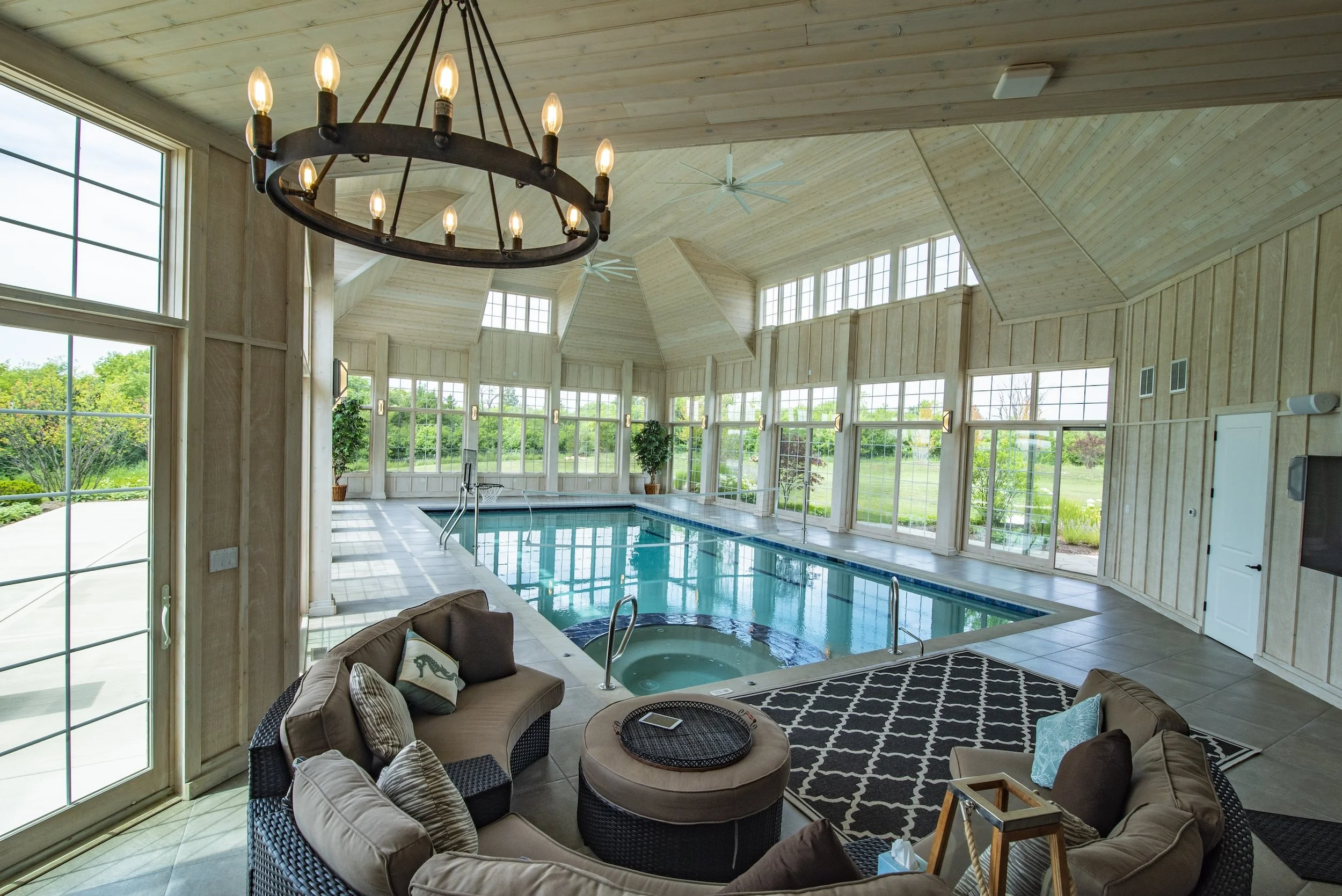 Indoor swimming pool with surrounding lounge area, large windows, and decorative chandelier.