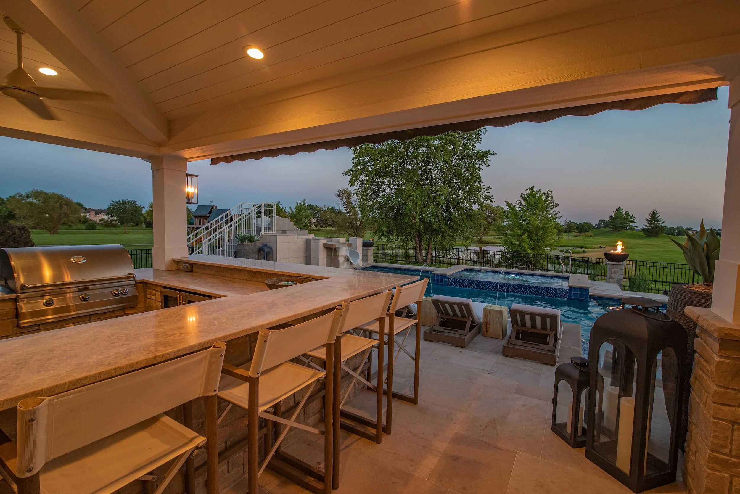 Outdoor kitchen with grill, bar seating, and a view of a swimming pool and landscaped yard in the background.