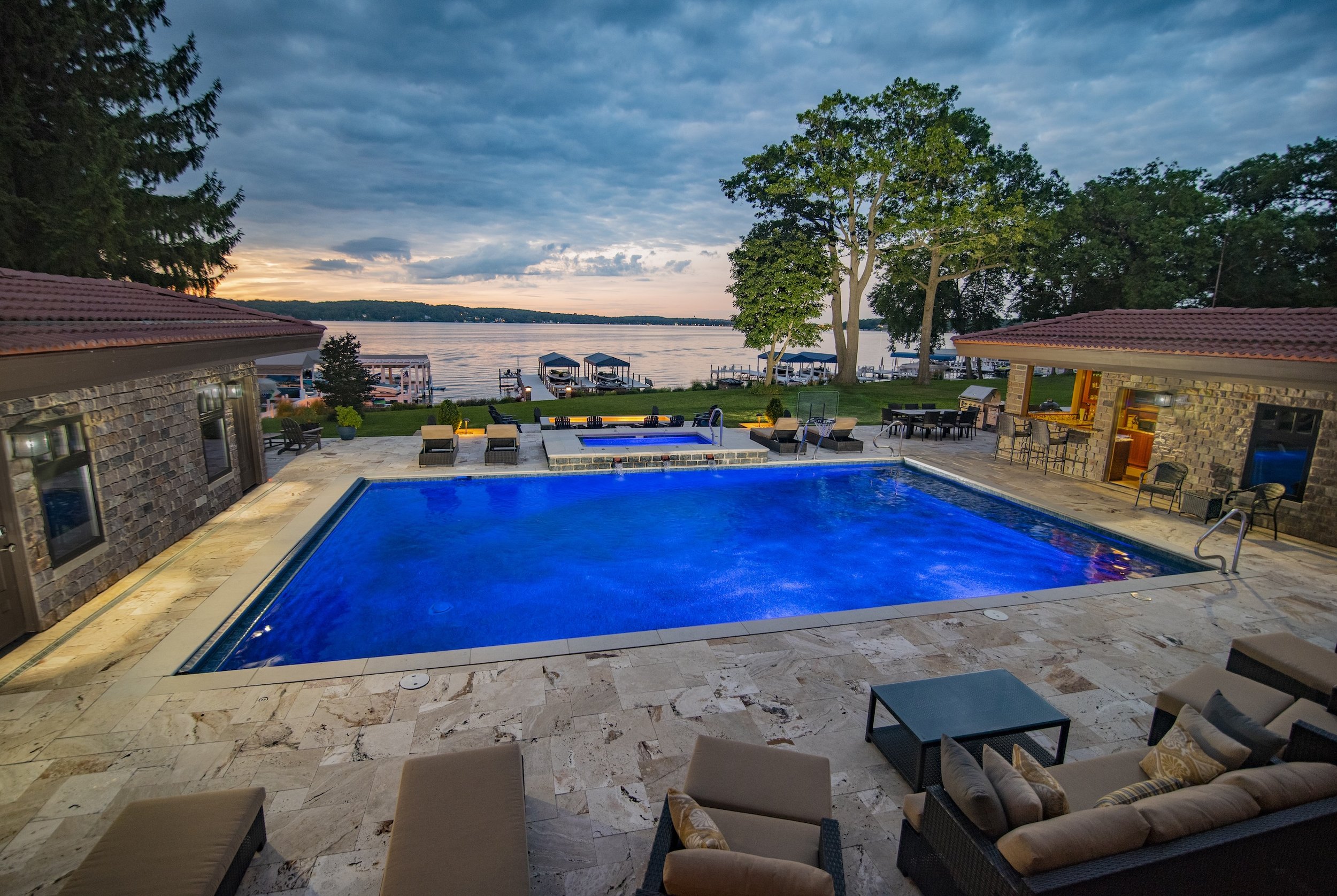 Luxury backyard with a swimming pool, loungers, outdoor furniture, and a view of a lake with docks and boats at sunset.