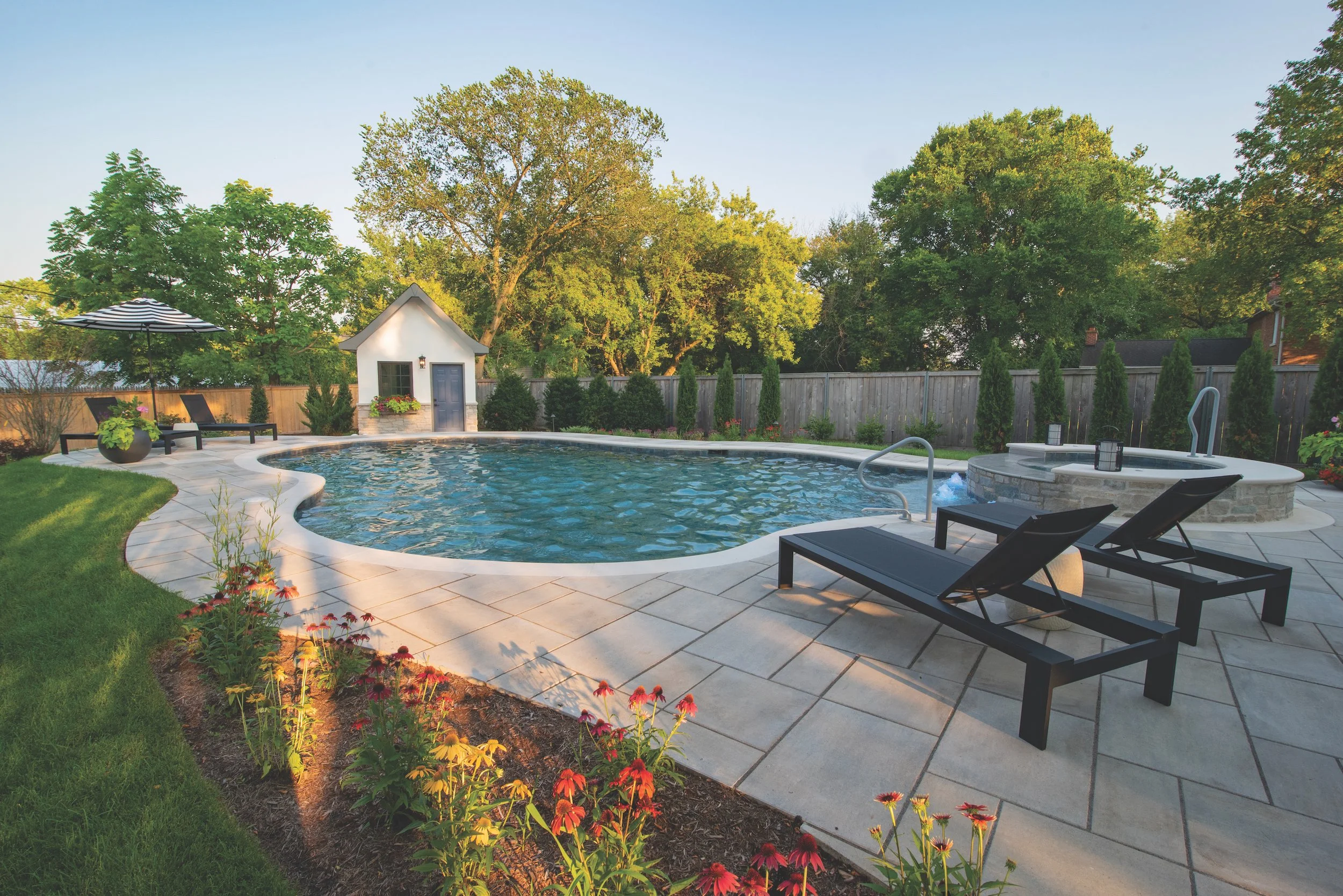 Backyard with swimming pool, spa, chaise lounges, landscaped garden, trees, and a small pool house.