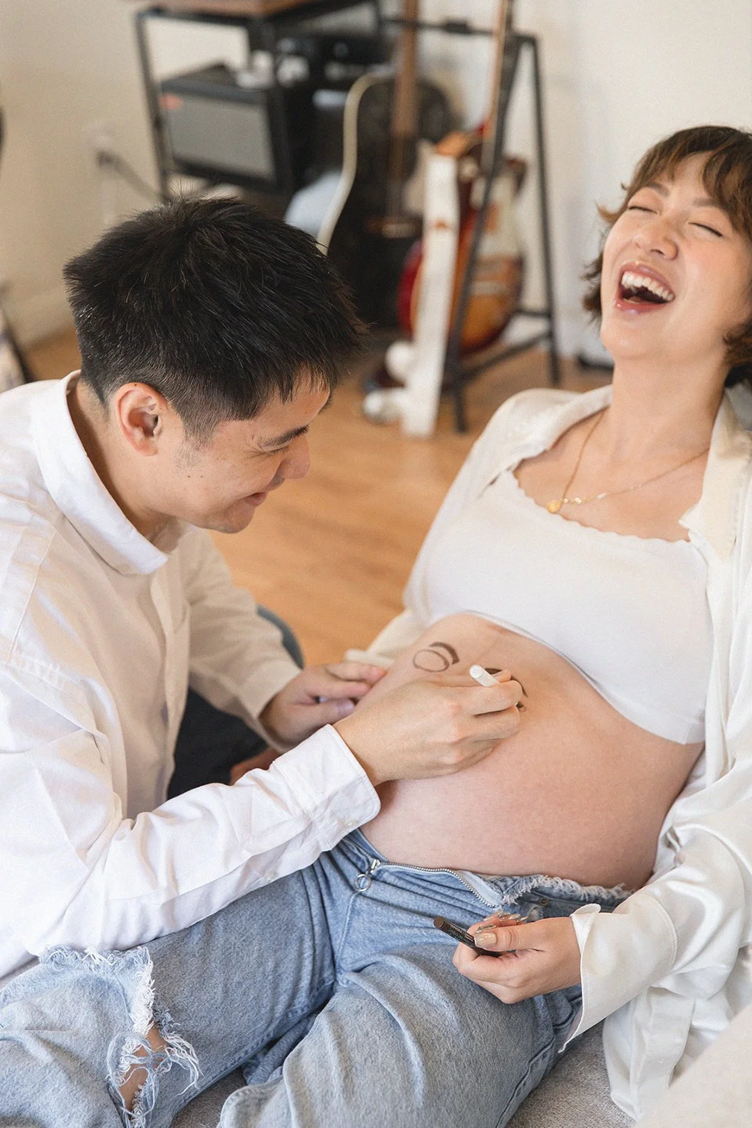 A man is drawing a heart on a pregnant woman's belly, and they are both laughing and happy.