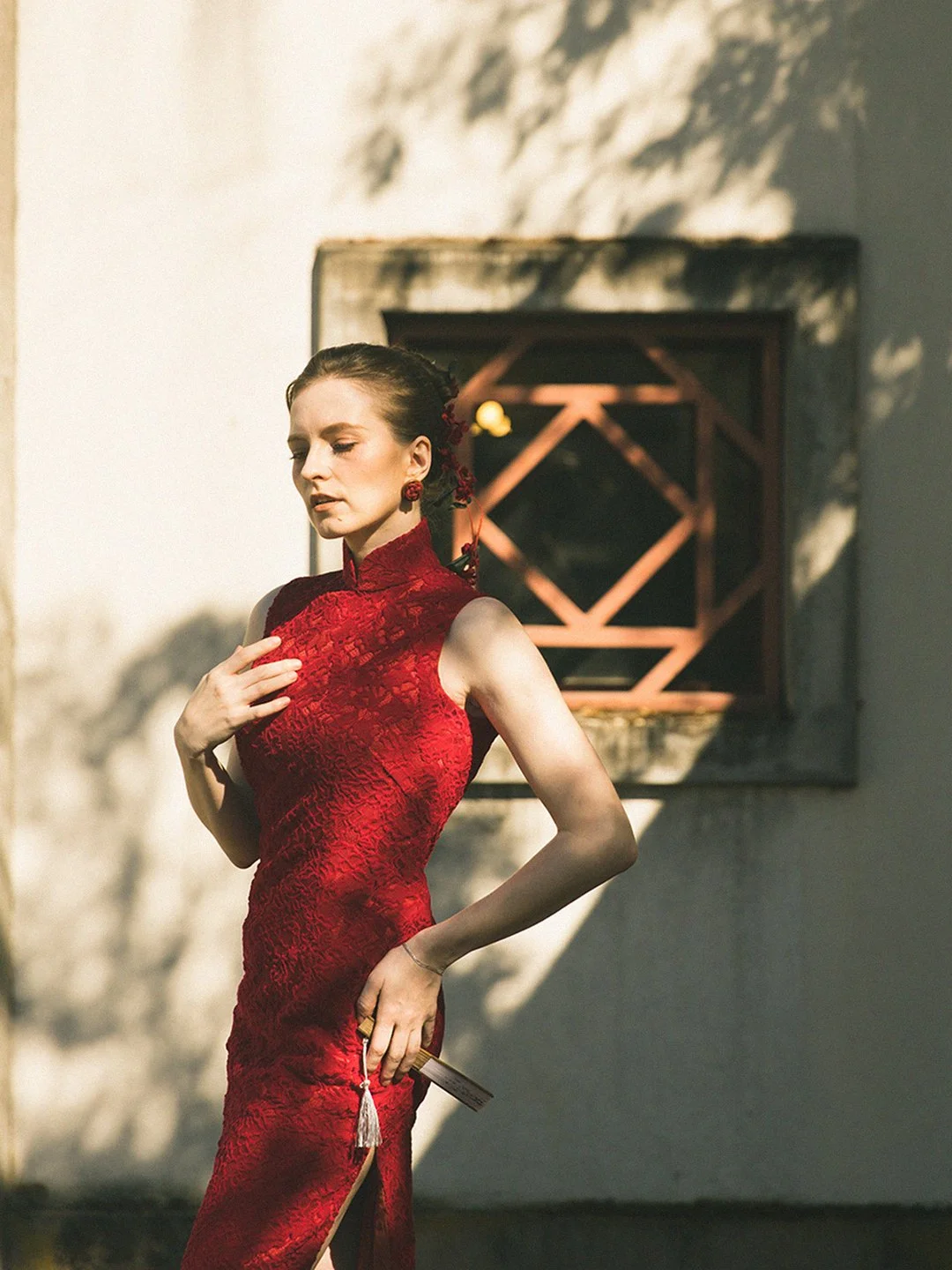 A woman in a red cheongsam dress standing outdoors with her eyes closed, holding a fan behind her back, in front of a white wall with a decorative wooden window.