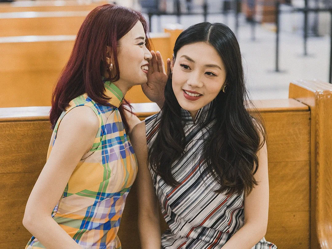 Two women are sitting close to each other, one whispering into the other's ear in what appears to be a library or study area.