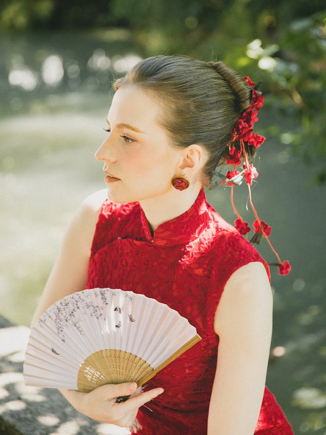A woman in a red traditional Chinese dress holding a fan, with her hair styled in an updo decorated with red flowers, standing outdoors near water.