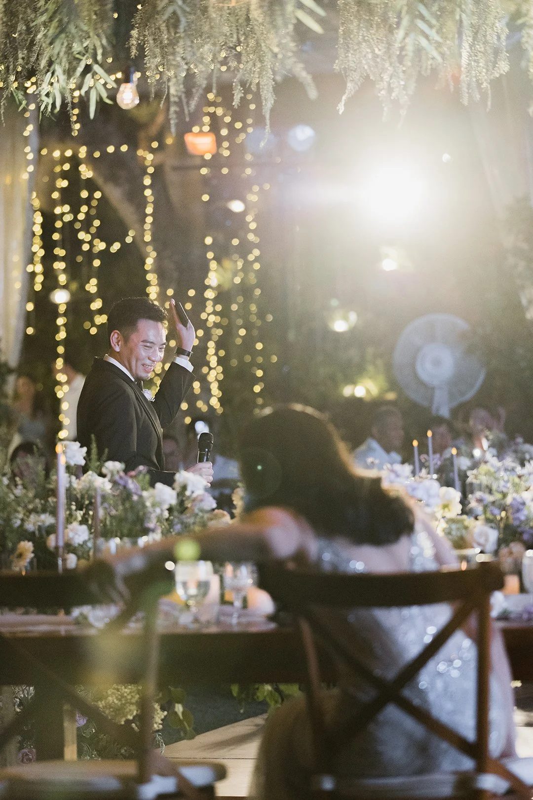 A man in a black tuxedo giving a speech at a wedding reception, standing behind a decorated table with flowers, surrounded by wedding guests, with fairy lights and hanging decorations in the background.