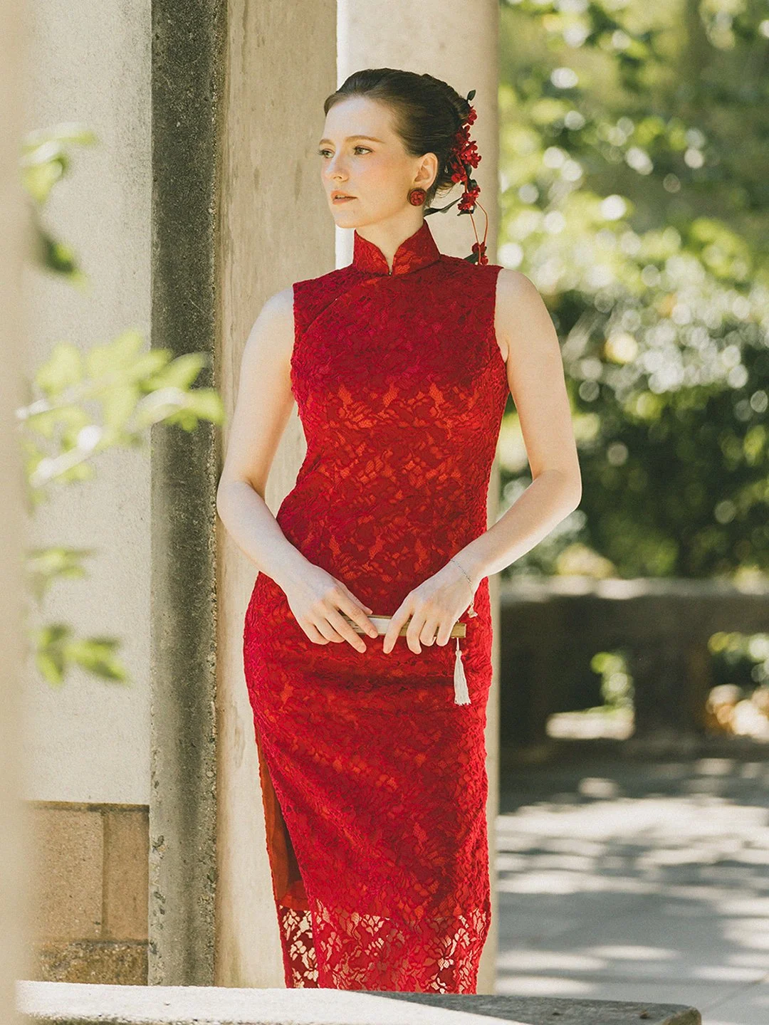 A woman in a red lace cheongsam dress standing outdoors near a stone pillar, holding a clutch, with trees and sunlight in the background.