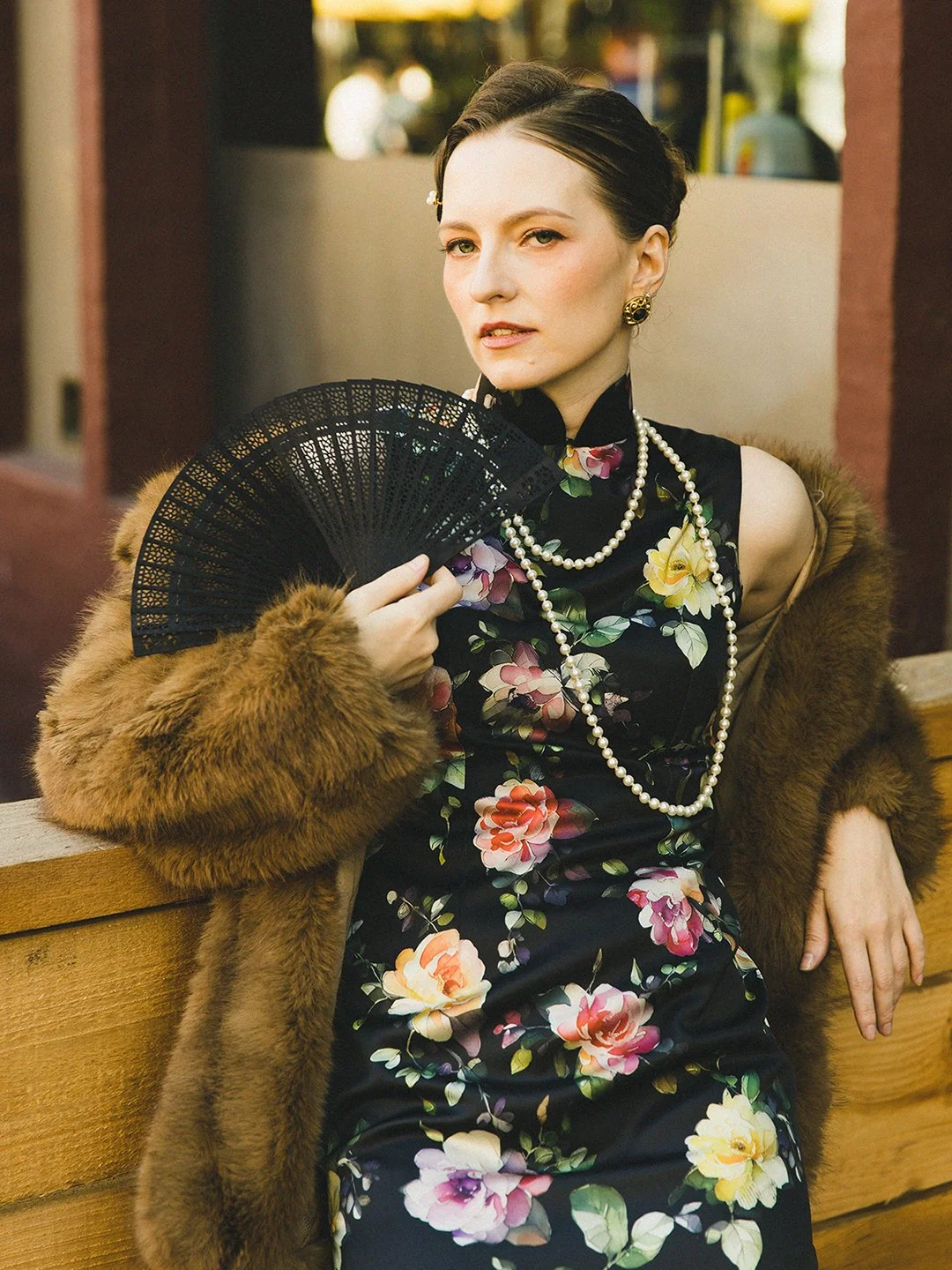 Woman sitting on a bench wearing a floral dress, pearl necklaces, earrings, a fur coat, and holding a black fan.