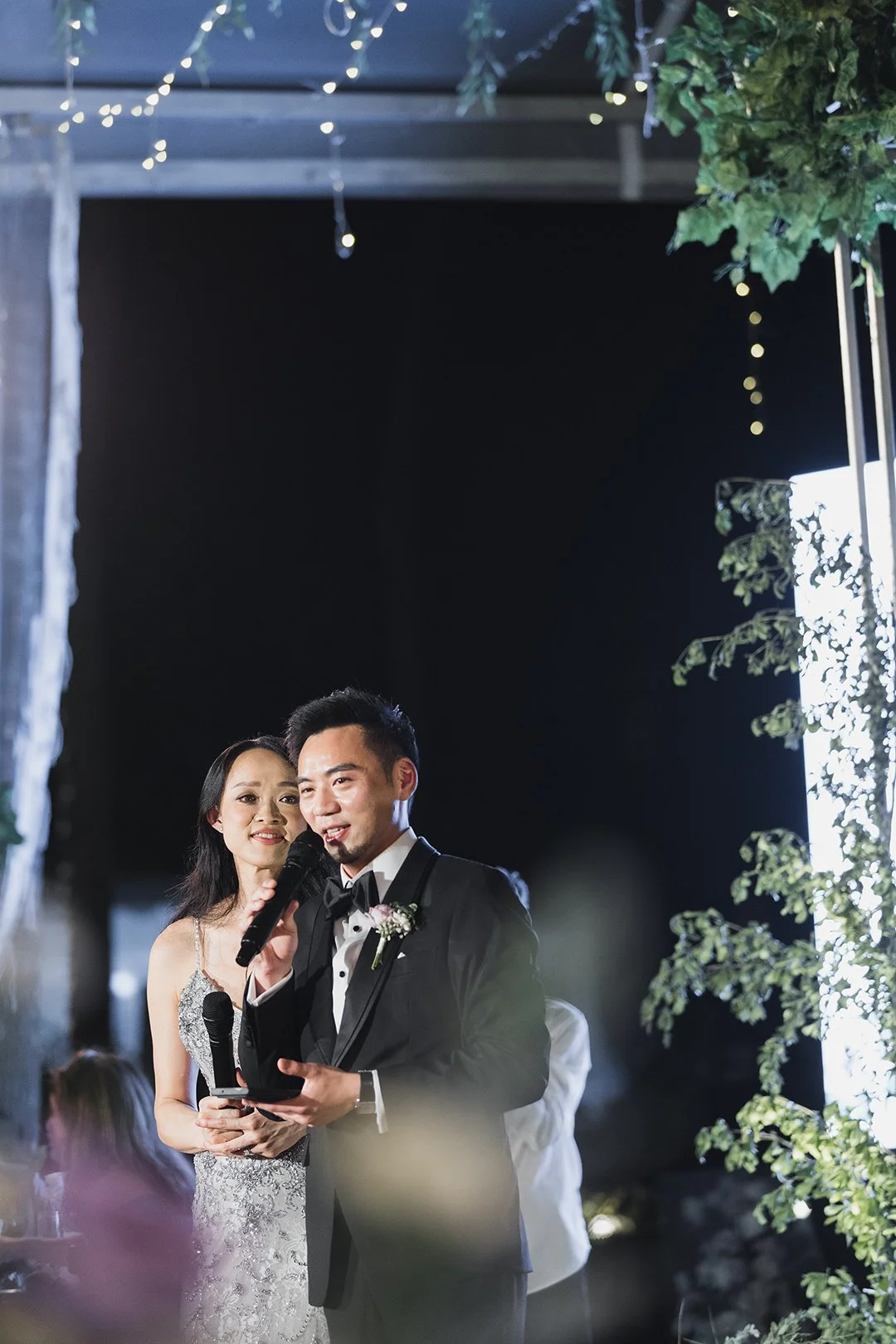 A man and a woman, dressed in formal wedding attire, stand together at a wedding reception. The man is speaking into a microphone while holding a small box. The woman looks on, smiling softly, in front of dark background decorations and string lights