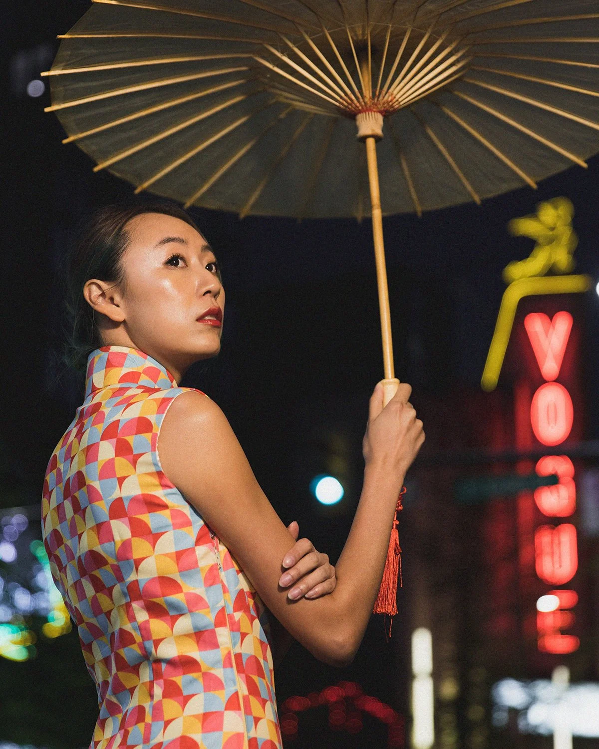 A woman standing outdoors at night under a traditional umbrella, wearing a colorful sleeveless dress with an abstract pattern, holding the umbrella with one hand, with illuminated neon signs in the background.