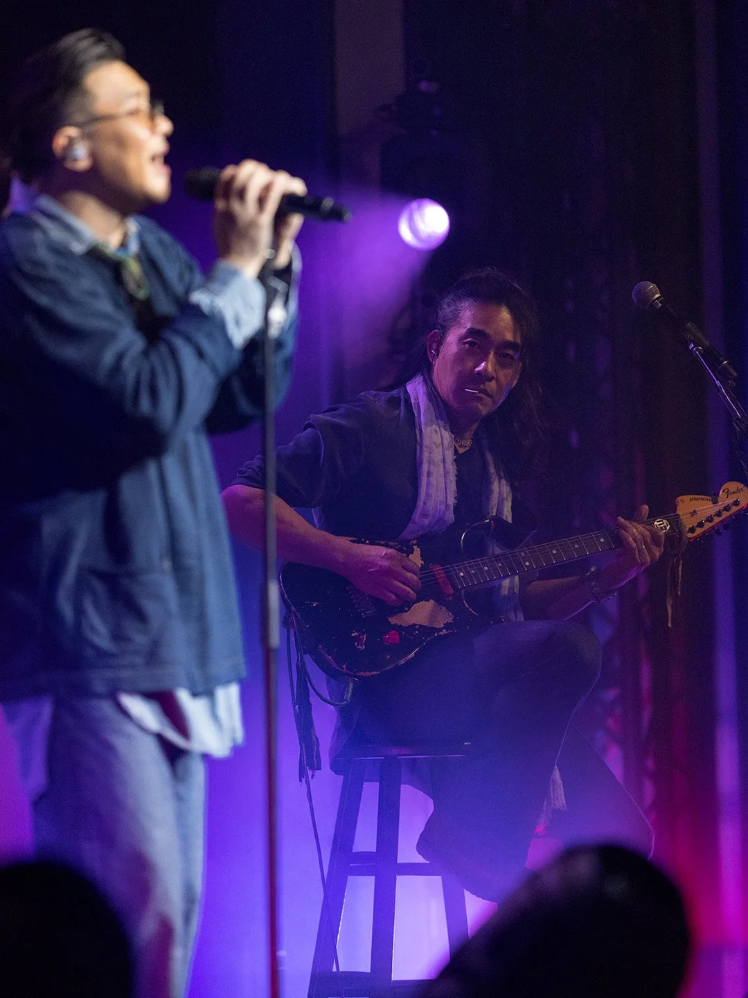 Musicians performing on stage with purple lighting, one person singing into a microphone, another playing an electric guitar.