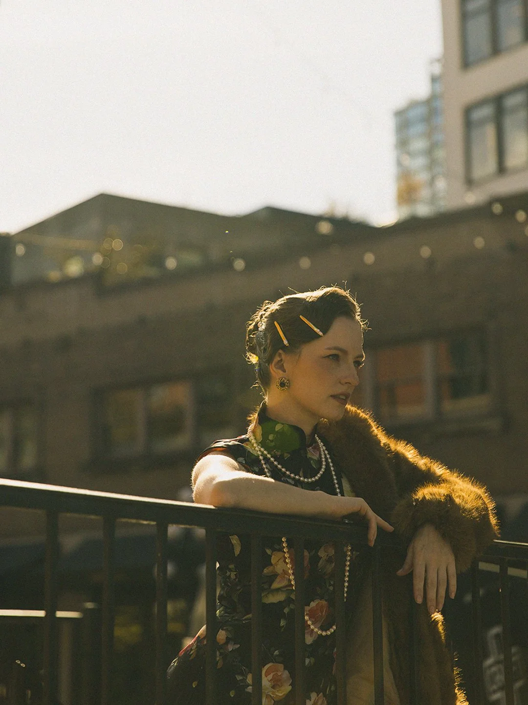 A woman with short dark hair, adorned with hair clips, wearing vintage jewelry, a floral dress, and a fur coat, stands on a bridge or balcony, leaning on a railing with a cityscape and buildings in the background, bathed in sunlight.