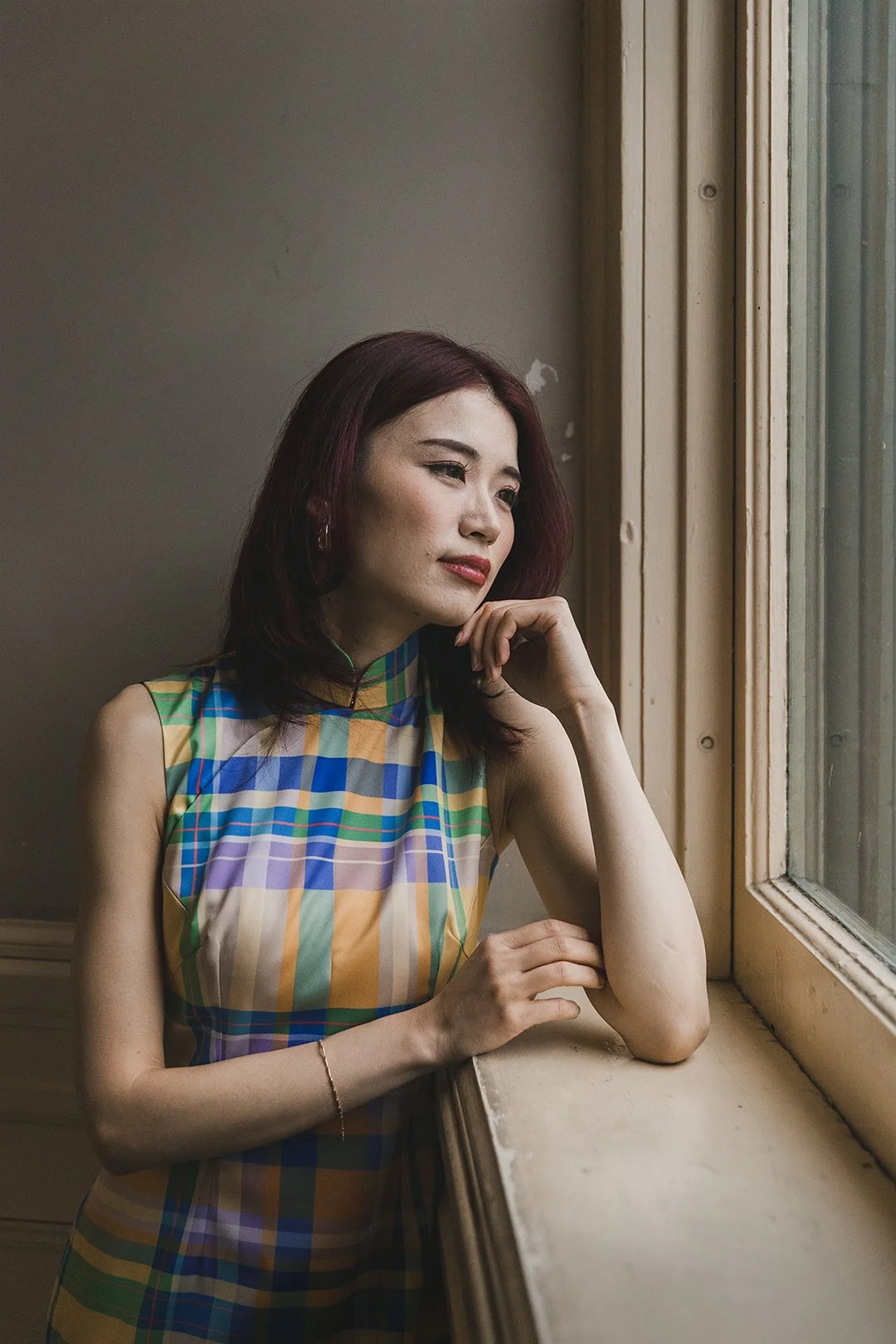 A woman with shoulder-length dark red hair wearing a colorful plaid sleeveless dress, gazing thoughtfully out a window.