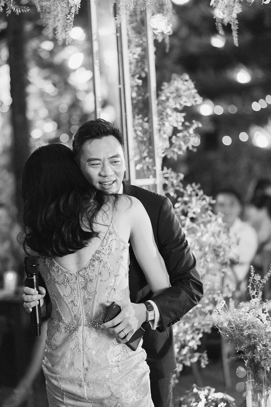 A man in a suit and a woman in an embellished dress embrace at a celebration, with blurred people and festive decorations in the background.