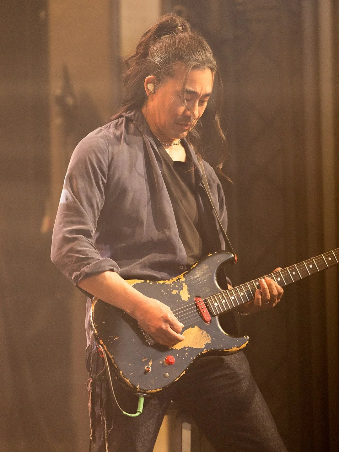 A man playing an electric guitar with worn paint on it, wearing a dark jacket and black T-shirt, during a performance.