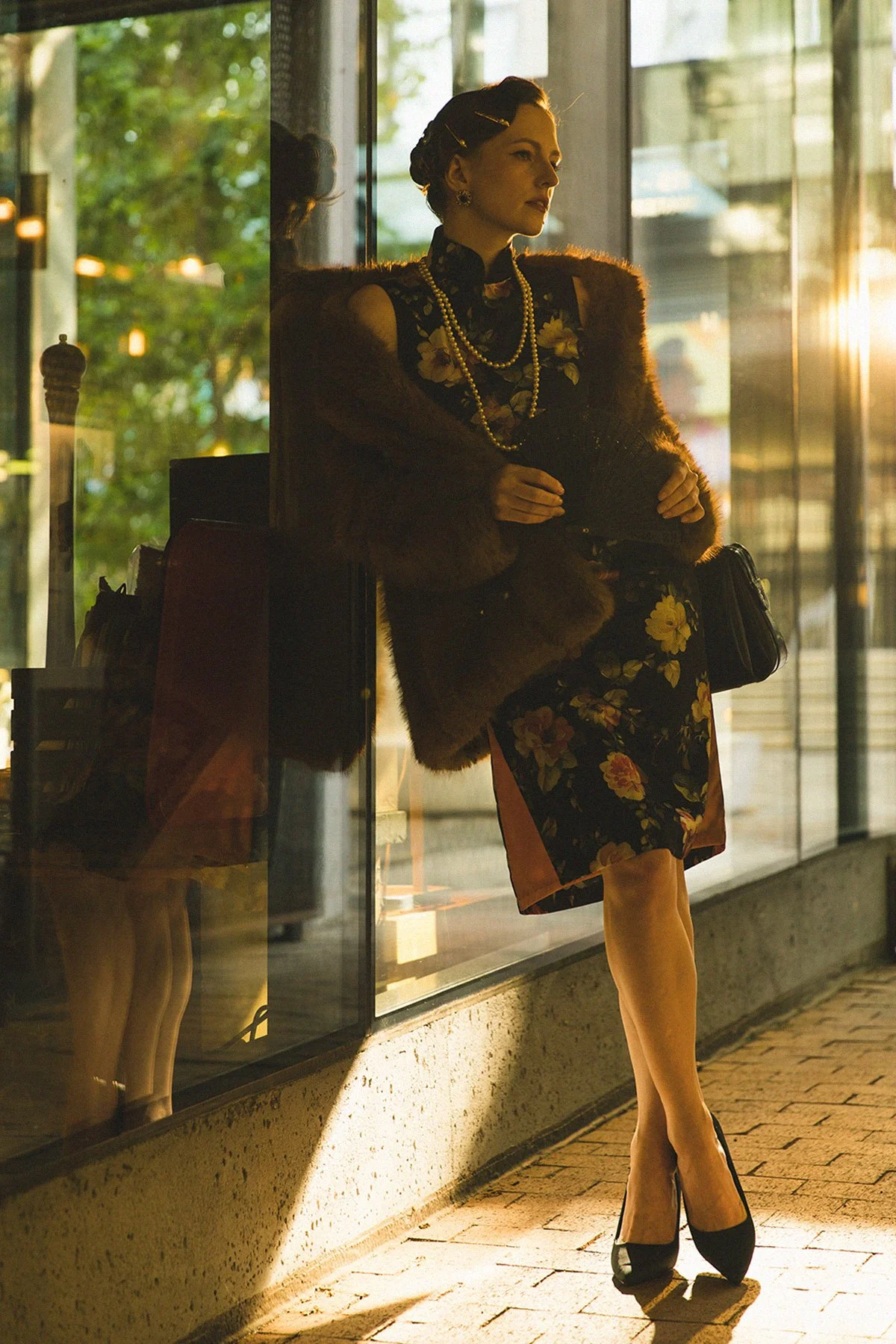 A woman standing outside a glass storefront during sunset, dressed in floral prints, high heels, and jewelry, with a fur coat draped over her shoulders.