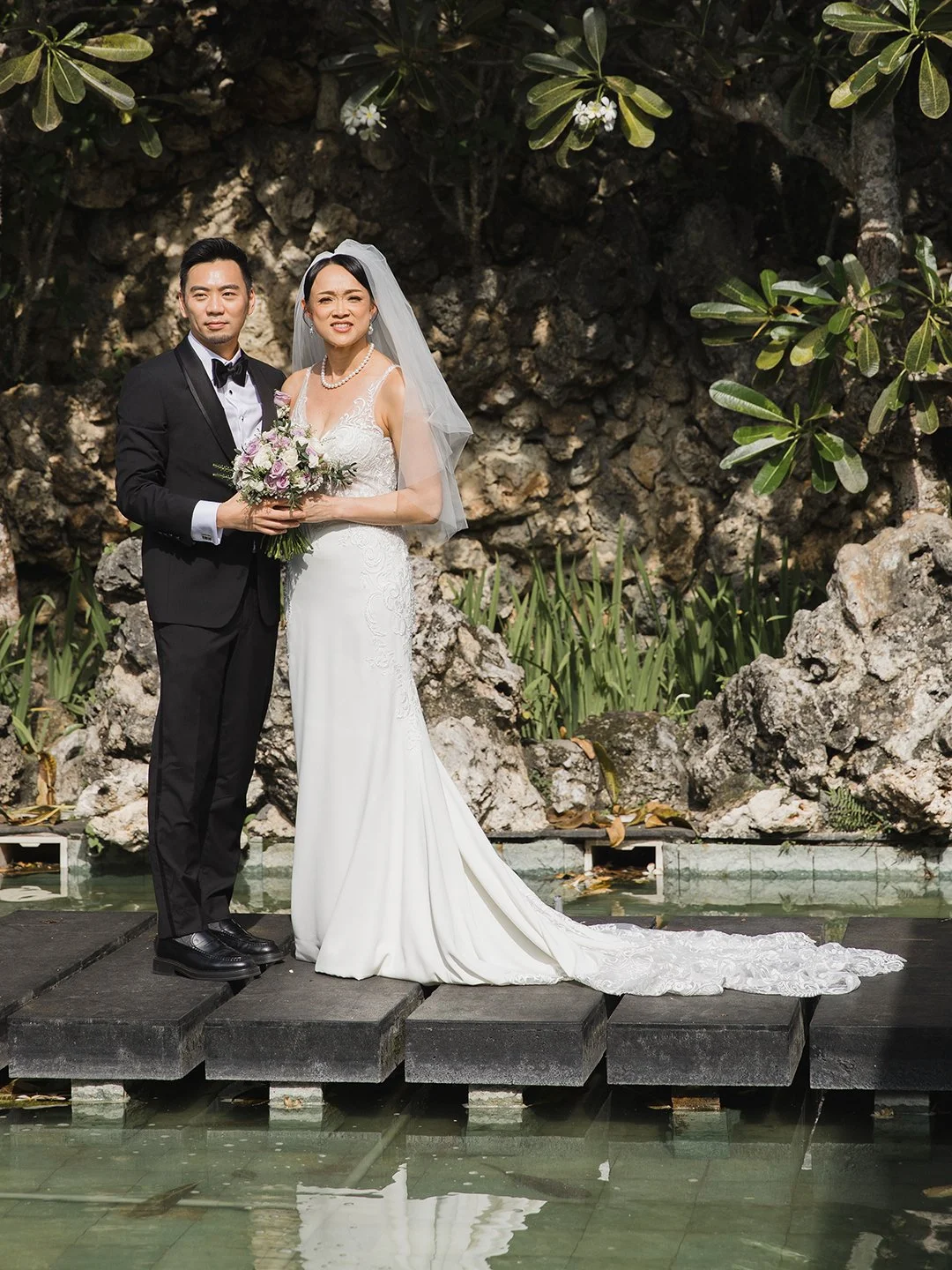 A bride and groom standing on a wooden platform in front of a rocky garden with greenery, posing for a wedding photo.