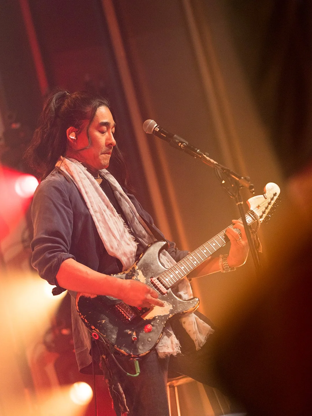 A musician playing an electric guitar on stage, with long black hair, wearing a dark shirt, a light scarf, and a wristwatch, illuminated by stage lights.