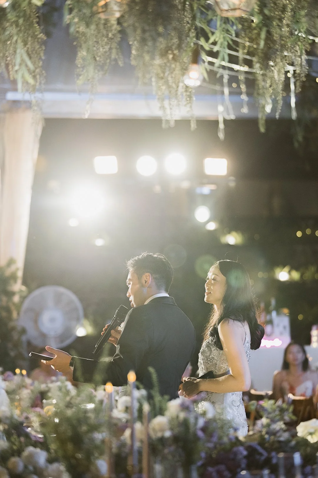 A couple during their wedding reception, with the groom holding a microphone and a piece of paper, and the bride smiling beside him, surrounded by flowers and bright lighting.