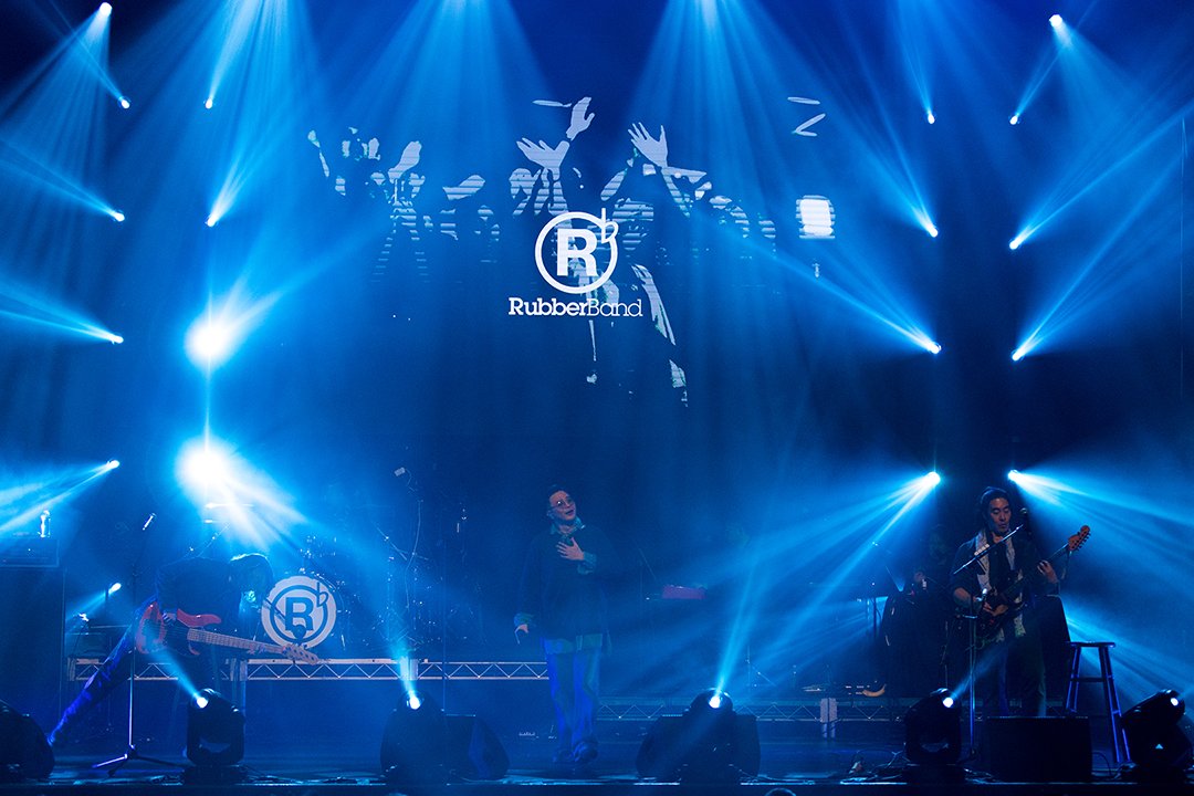 A live concert performance by RubberBand on stage with blue lighting, musical instruments, and a large screen displaying the band's logo.