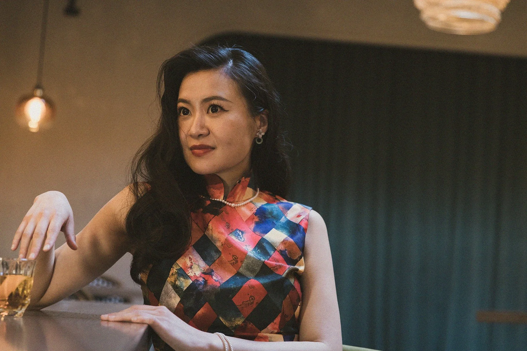 A woman with long black hair, wearing a colorful patterned sleeveless dress, pearl necklace, and earrings, sitting at a bar with a glass of drink in front of her, against a dimly lit background.