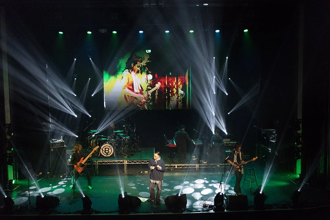 Stage with live band performing, large screen displaying a guitarist, with colorful lighting and spotlights.