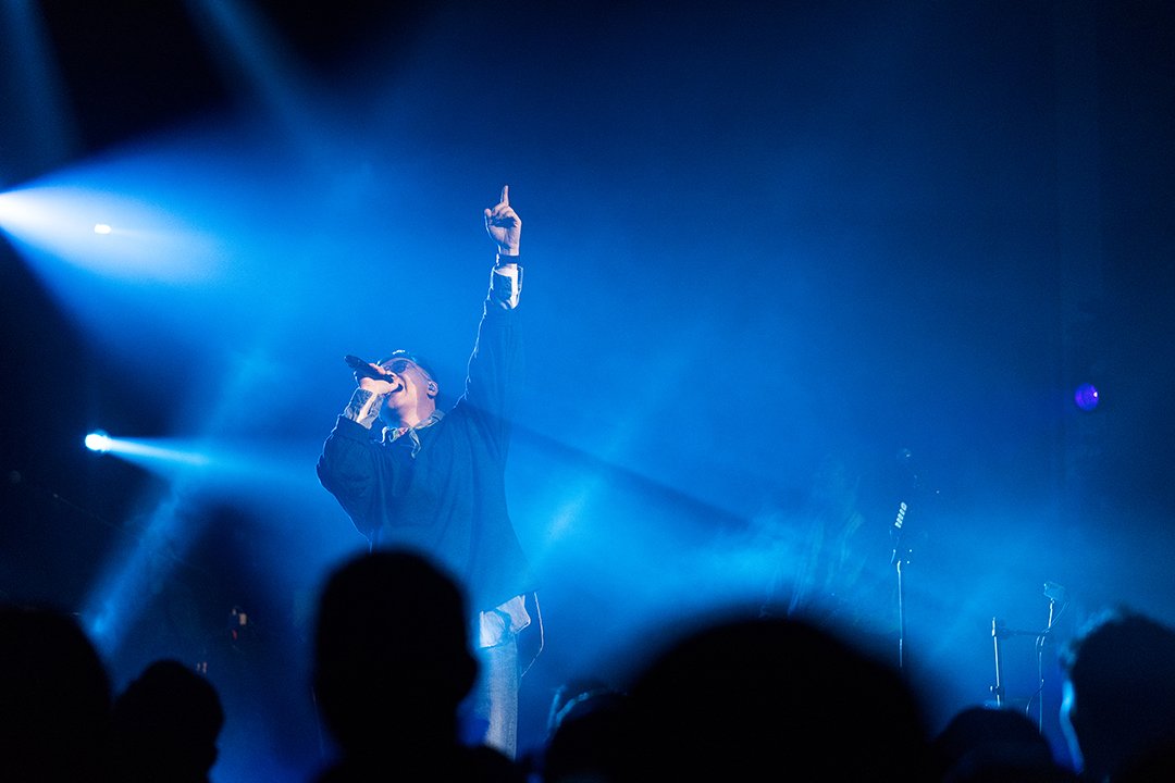 A singer performing on stage with blue lighting, holding a microphone, and raising one hand.