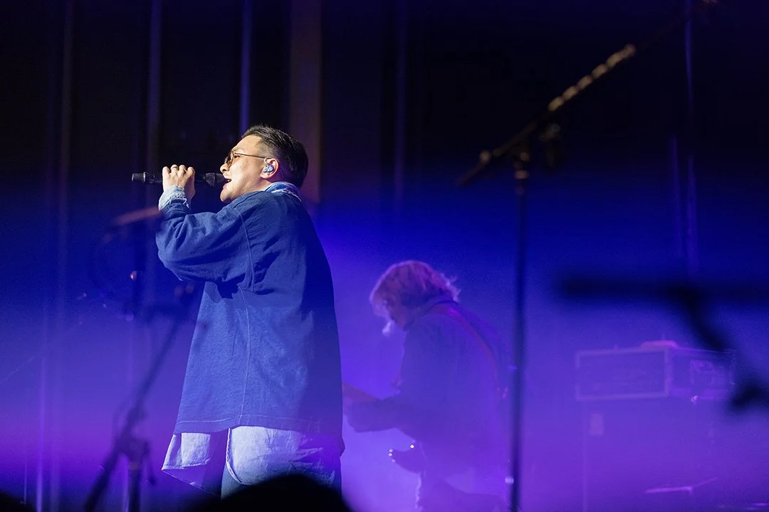Male singer in glasses and a blue jacket performing on stage with purple lighting, accompanied by a musician in the background.