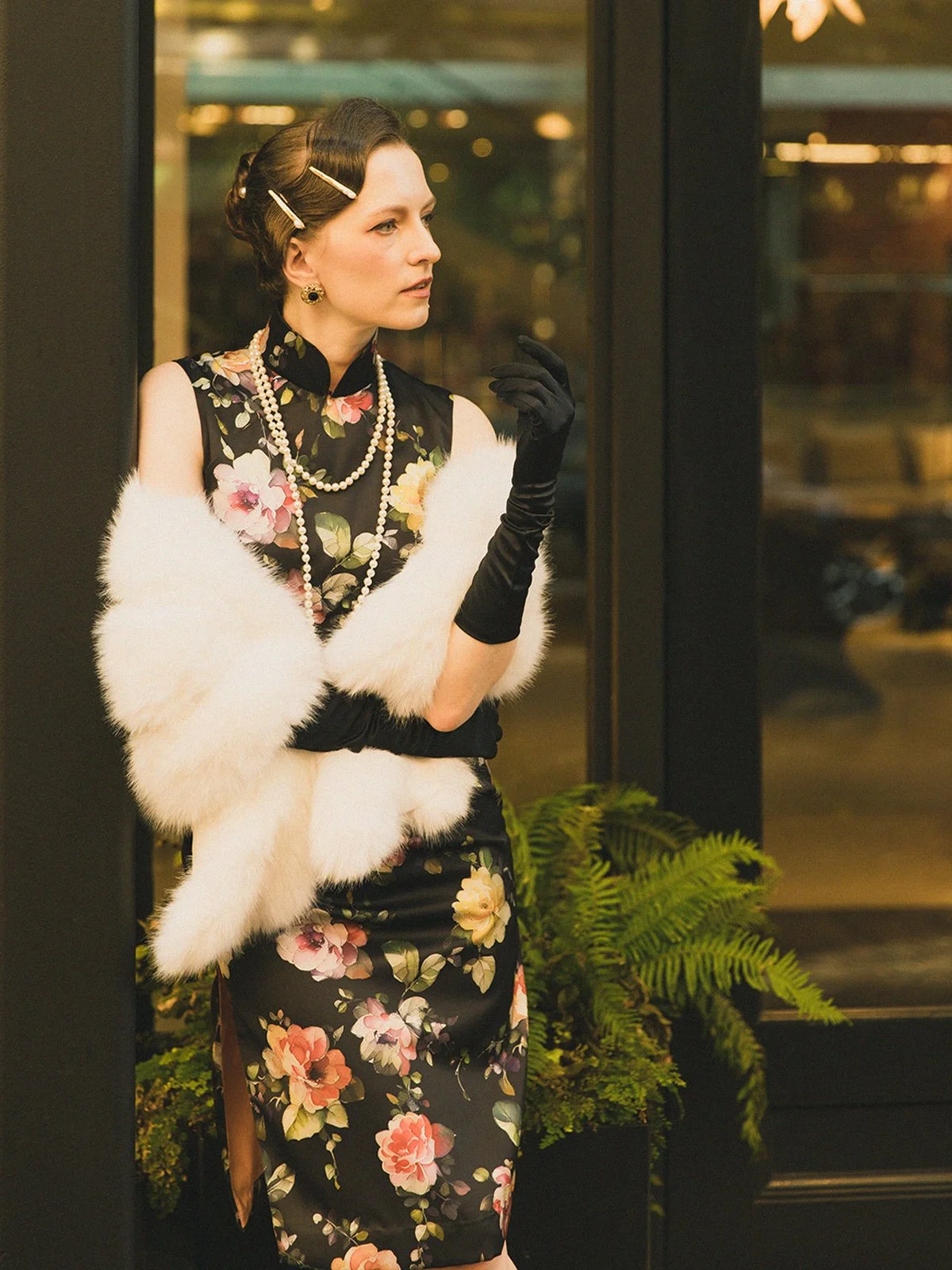 A woman in vintage-style attire standing by a glass door, wearing a floral dress, pearl necklaces, black gloves, and a white fur stole.