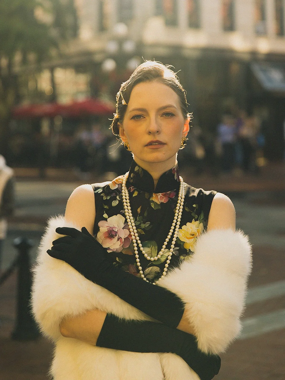 A woman in a floral dress with pearl necklaces, black gloves, and a white fur stole standing in an outdoor urban setting with buildings and people in the background.