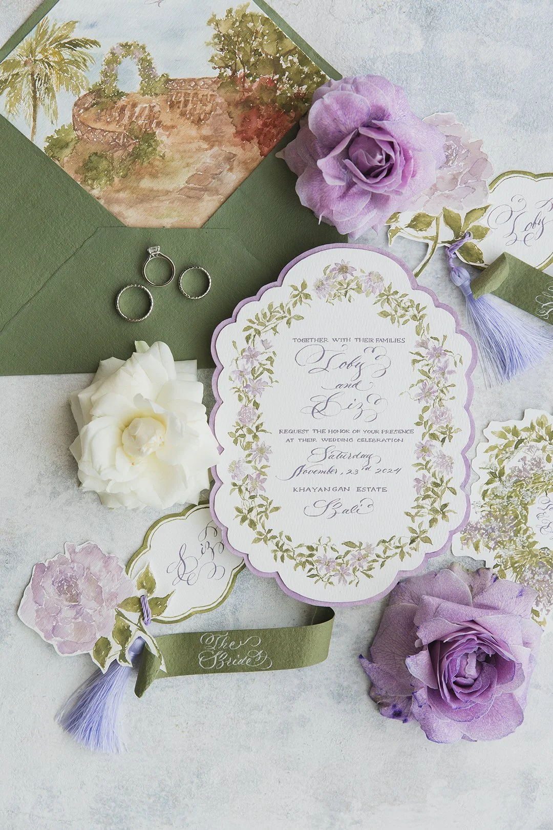 Wedding invitation set with floral motifs, purple and white roses, rings, envelope, and decorative paper items on a light-colored surface.