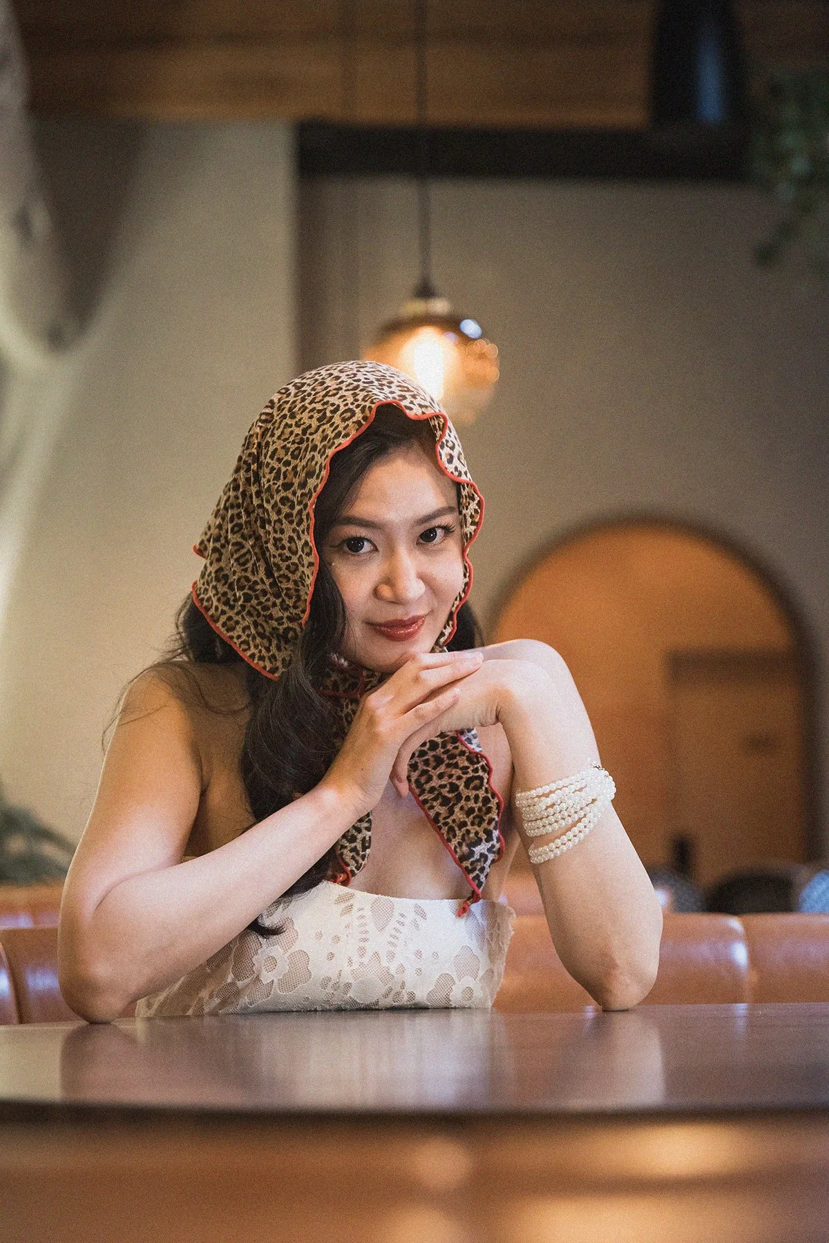 A woman with dark wavy hair wearing a leopard print scarf on her head, a white lace dress, and pearl bracelets, sitting at a table in a cozy restaurant with warm lighting and wooden accents.