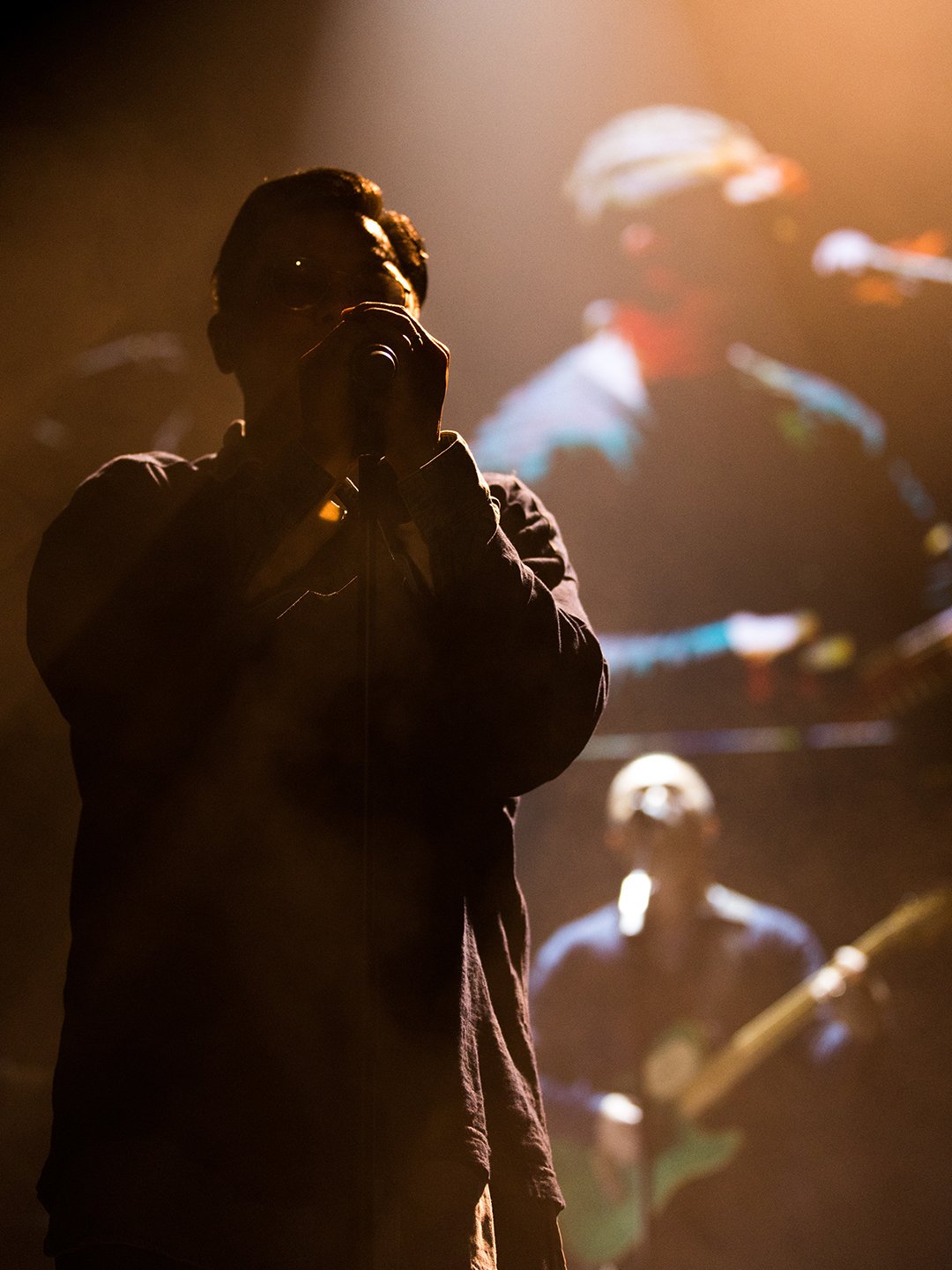 Silhouette of a singer holding a microphone on stage with a guitarist in the background, illuminated by warm stage lighting.