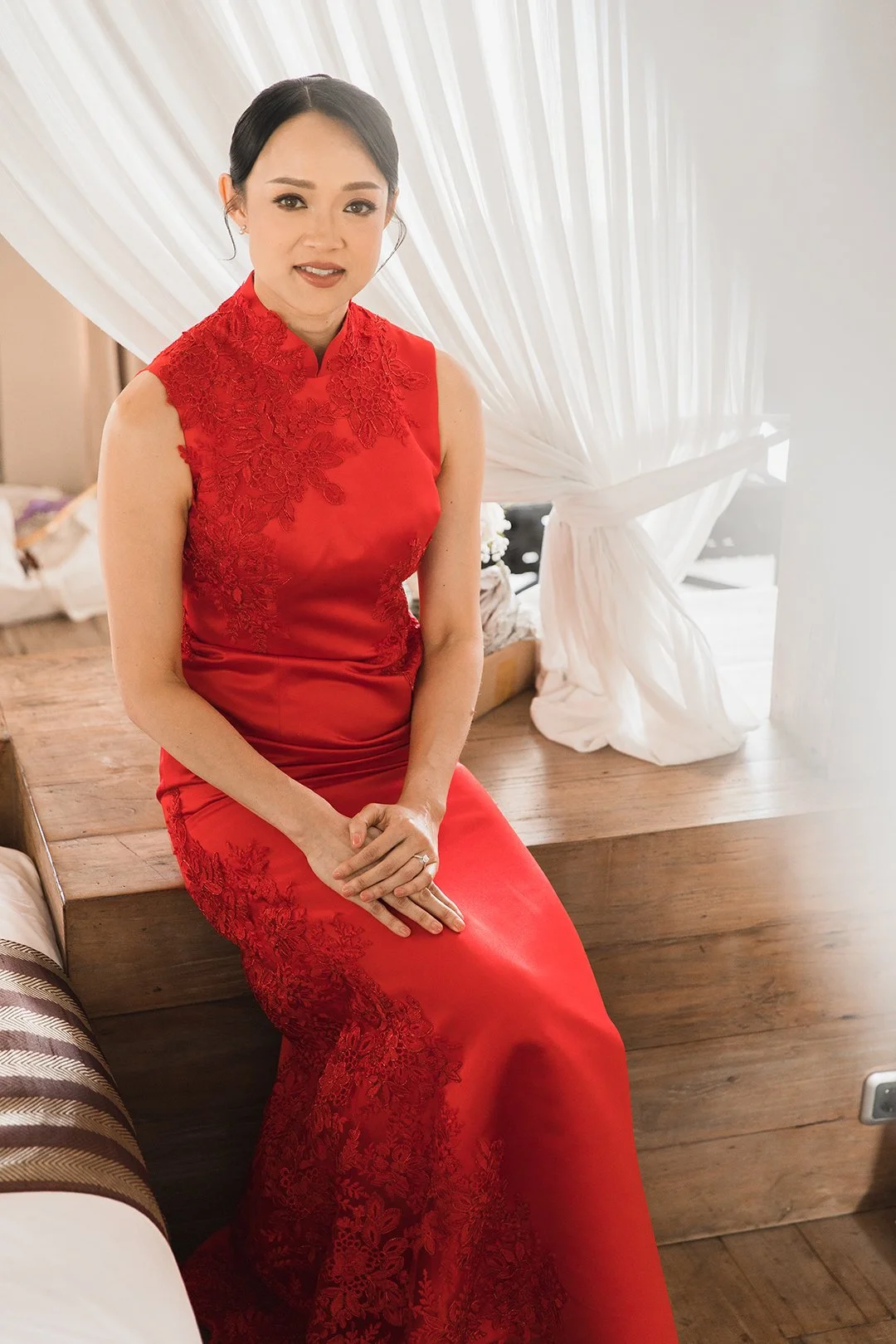 A woman in a red Chinese-style dress with lace details sitting on a wooden bench near a window with white curtains.