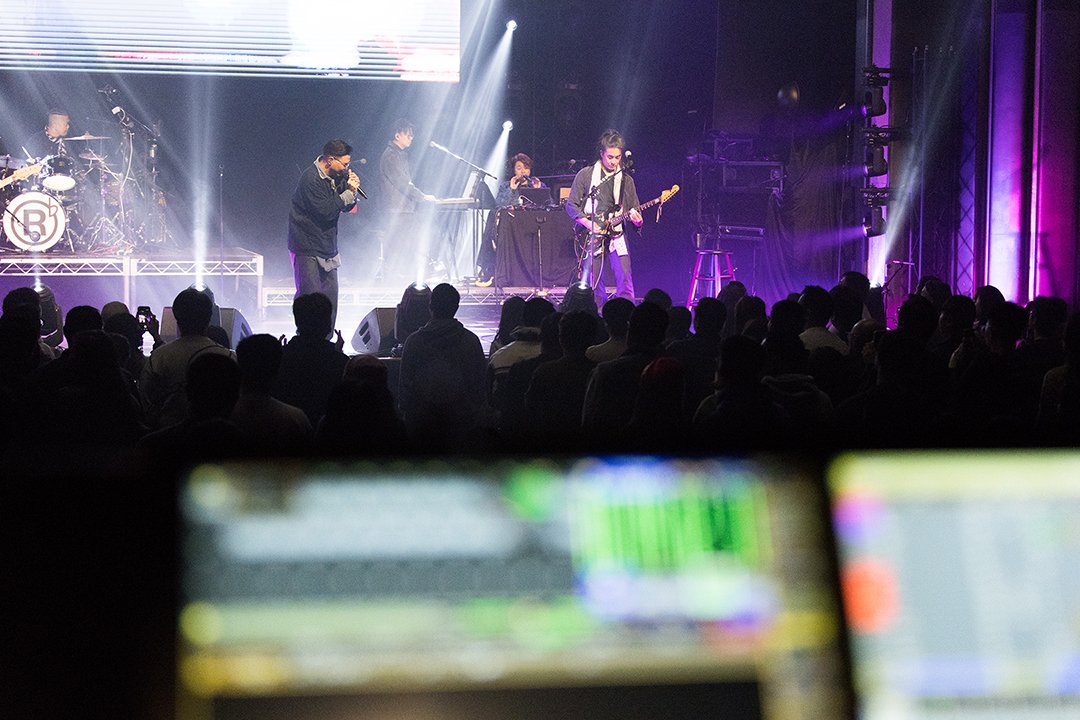Musicians perform on stage with audience watching, colorful stage lights and sound equipment visible.