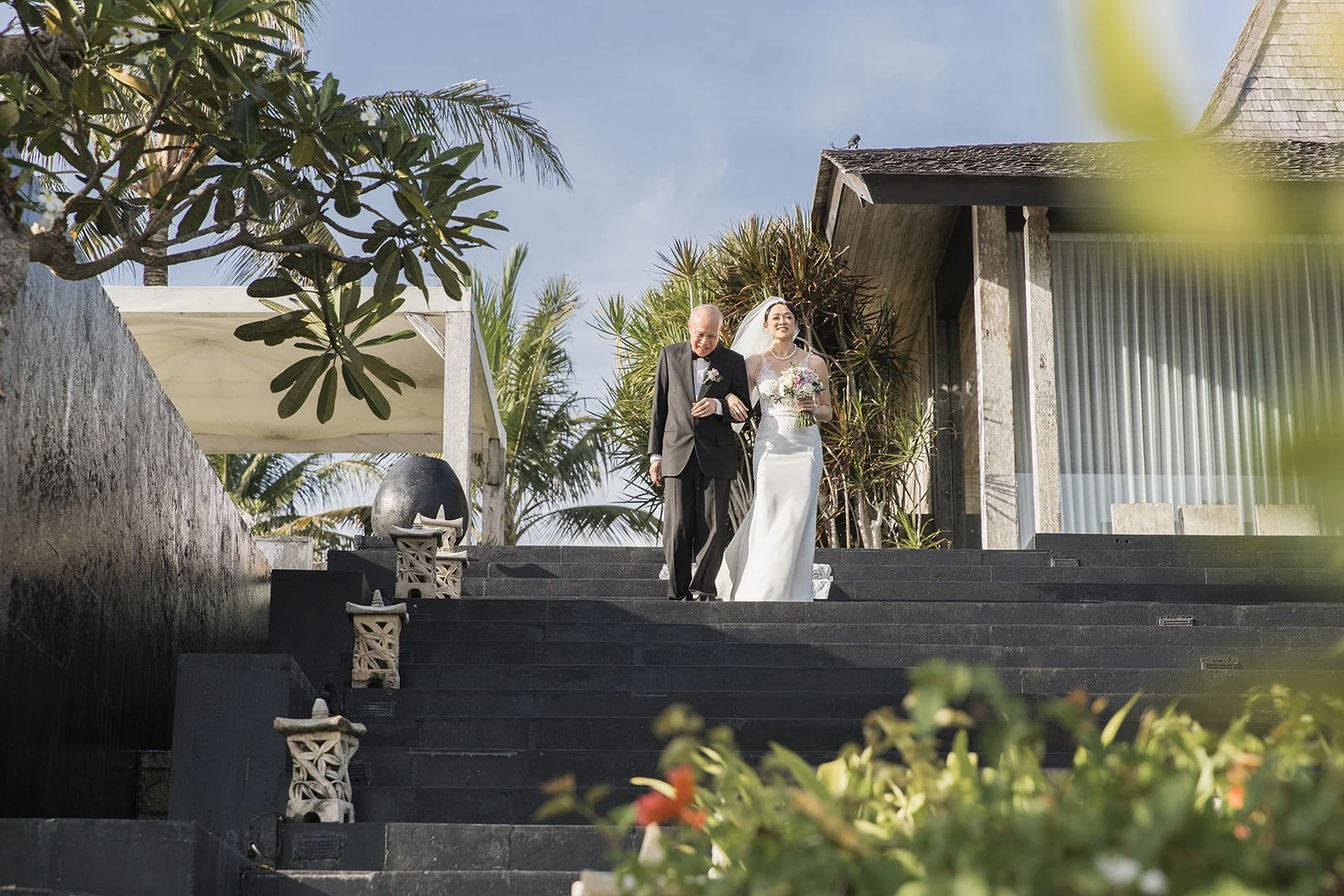 A bride and an older man walking down outdoor steps at a wedding venue with lush green plants and a modern building in the background, under a clear blue sky.