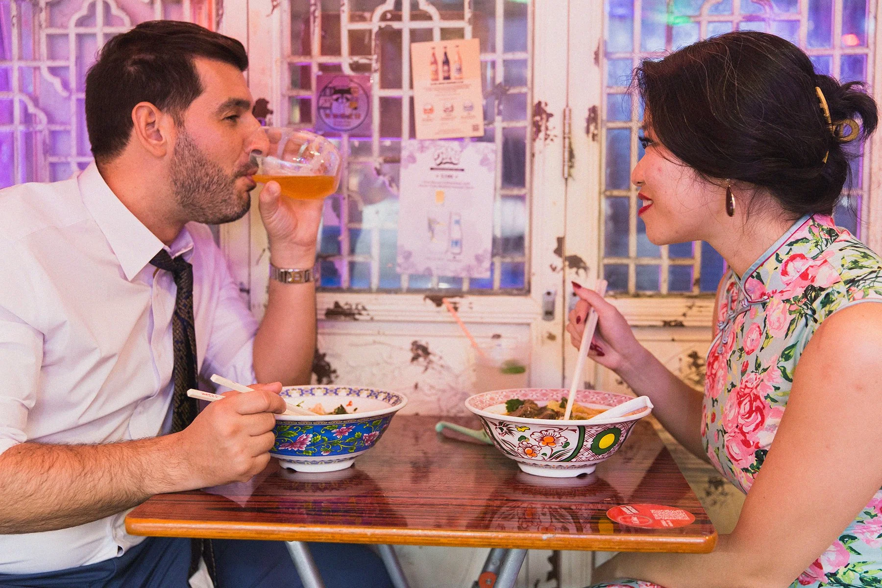 A man and woman are sitting at a wooden table in a restaurant, enjoying a meal together. The man is drinking a glass of beer, while the woman is smiling and eating from a bowl with chopsticks.