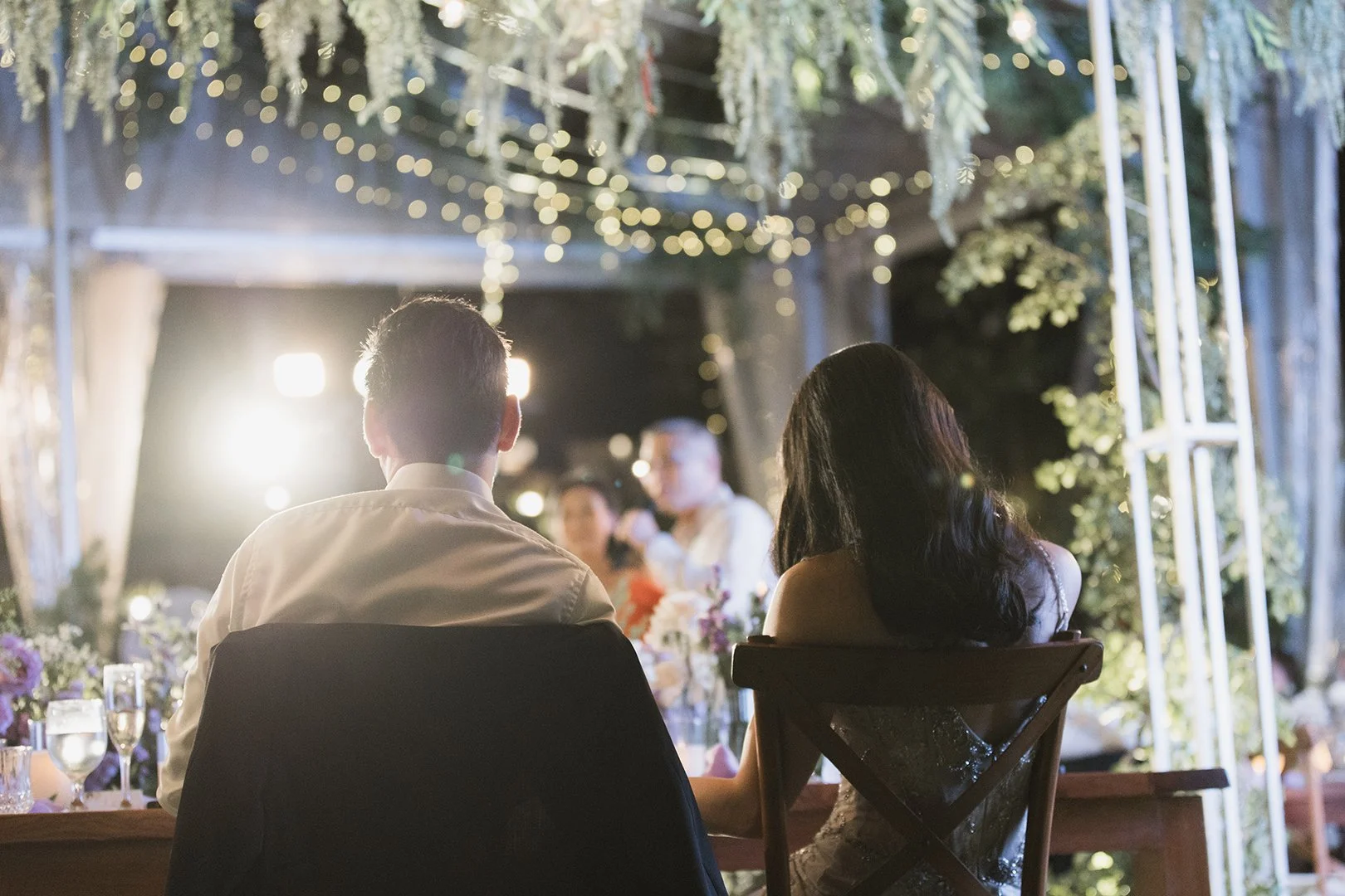Couple at a wedding reception sitting at a table with string lights and floral decorations behind them.
