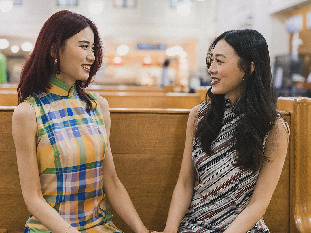 Two women sitting on a bench in a mall, smiling and holding hands. The woman on the left has red hair and wears a colorful, sleeveless dress with a checkered pattern. The woman on the right has black hair and wears a striped, sleeveless dress.