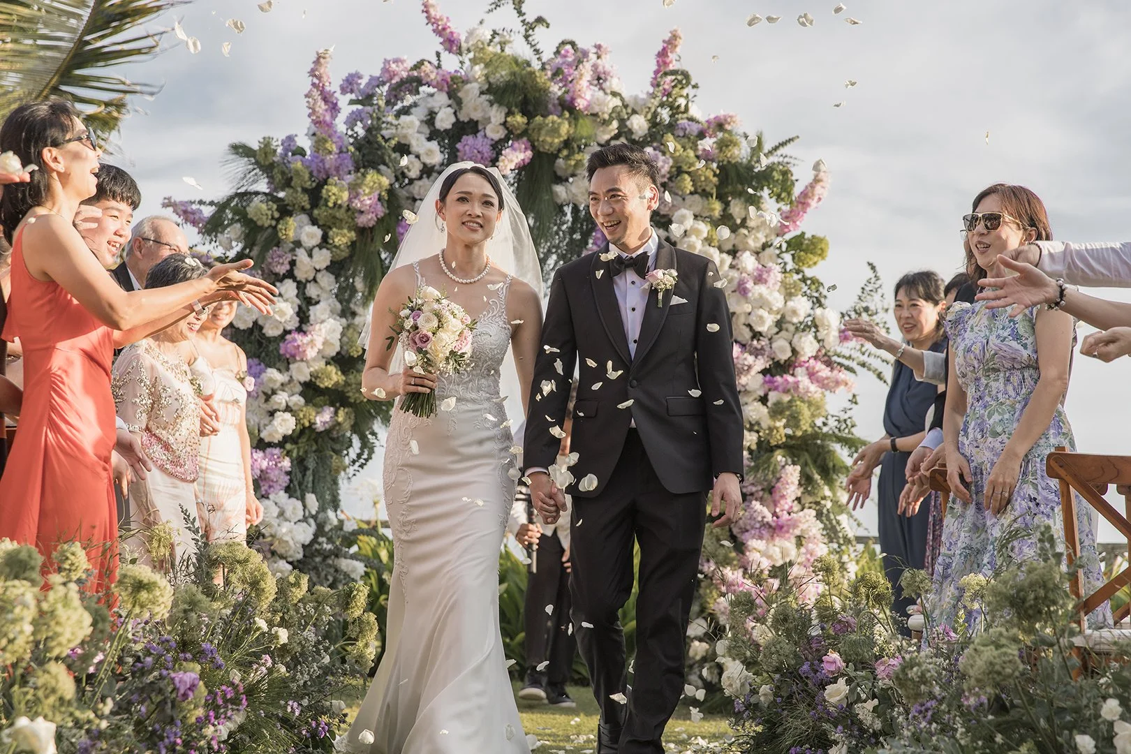 A bride and groom walking hand in hand down an outdoor wedding aisle, surrounded by friends and family cheering and throwing flower petals. The scene features a large floral arch in the background and bright, natural lighting.