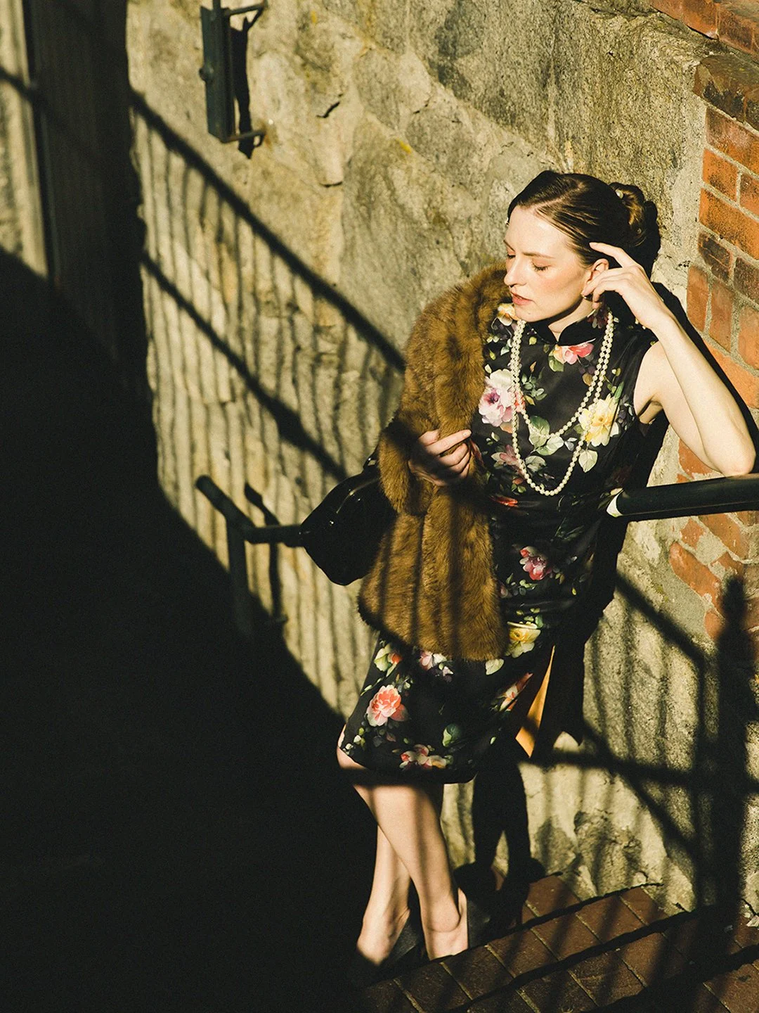 A woman with dark hair styled in an updo, wearing a black dress with a floral pattern, pearl necklace, holding a fur stole and a black purse, standing on a brick and stone stairway next to a brick and stone wall.