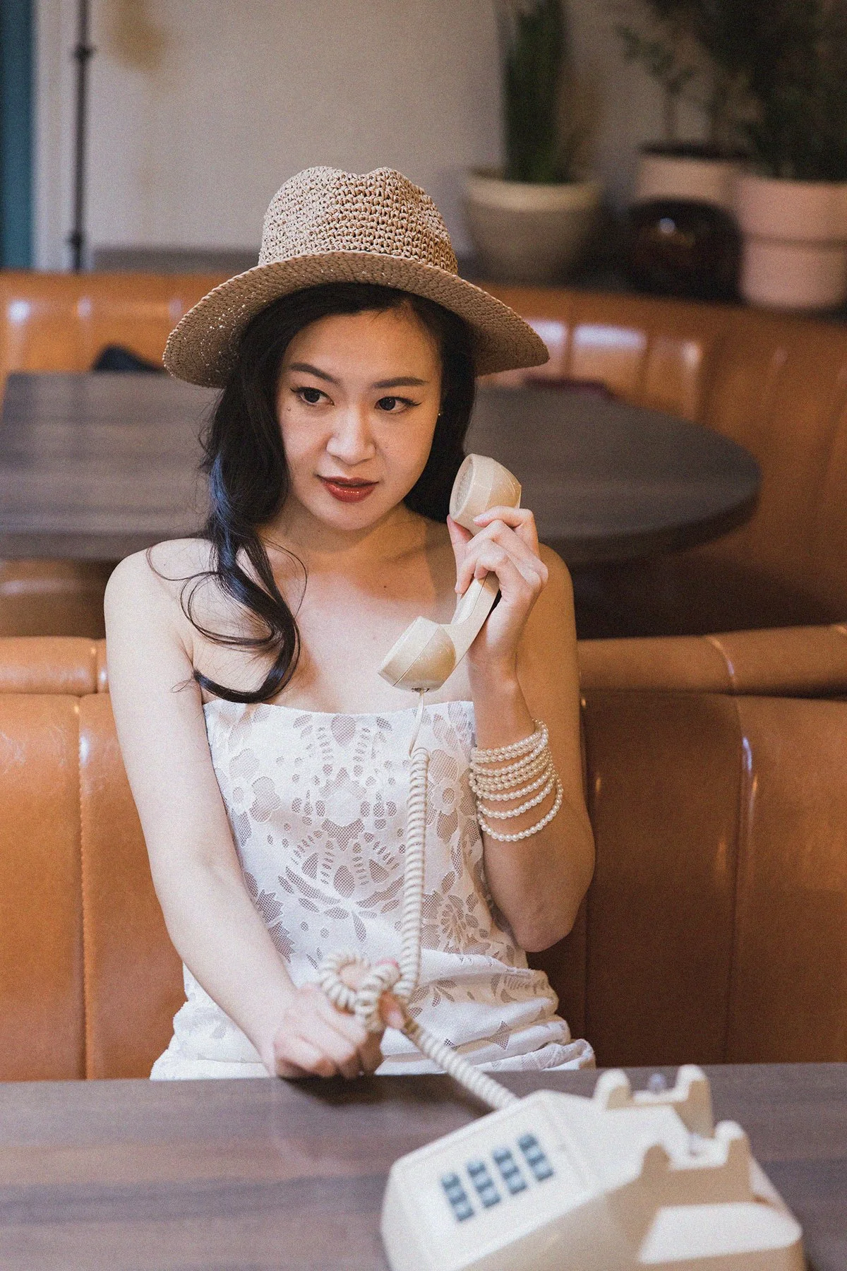A woman with dark hair wearing a straw hat, lace dress, and pearl bracelets, sitting in a vintage-style cafe, holding a beige rotary phone to her ear.