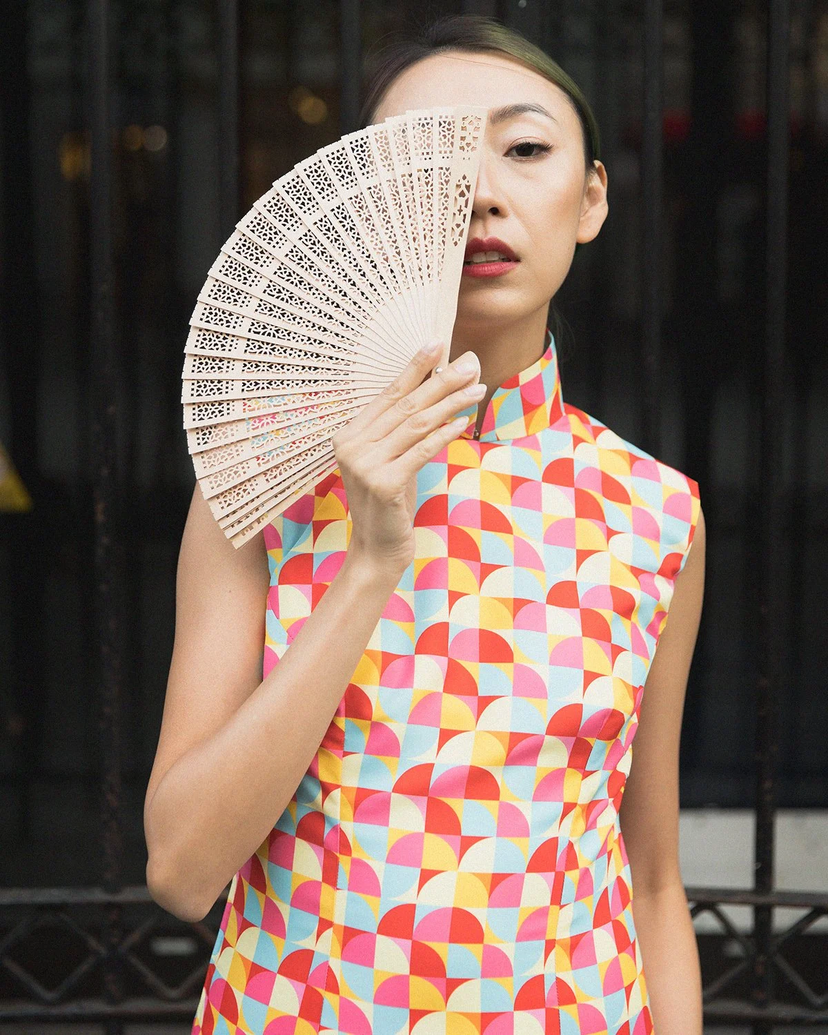 A woman with light skin and dark hair holds a decorative pink fan in front of her face, wearing a colorful sleeveless dress with a geometric pattern, standing in front of a dark background.
